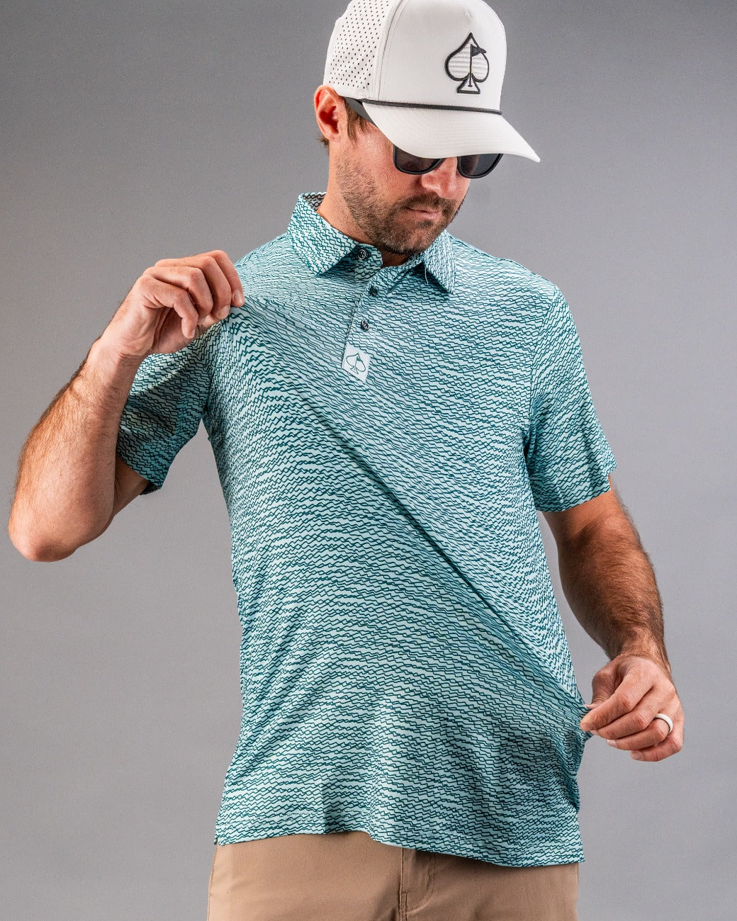 Man adjusting a teal polo shirt with a squiggle pattern, wearing sunglasses and a cap, showcasing bold golf apparel.