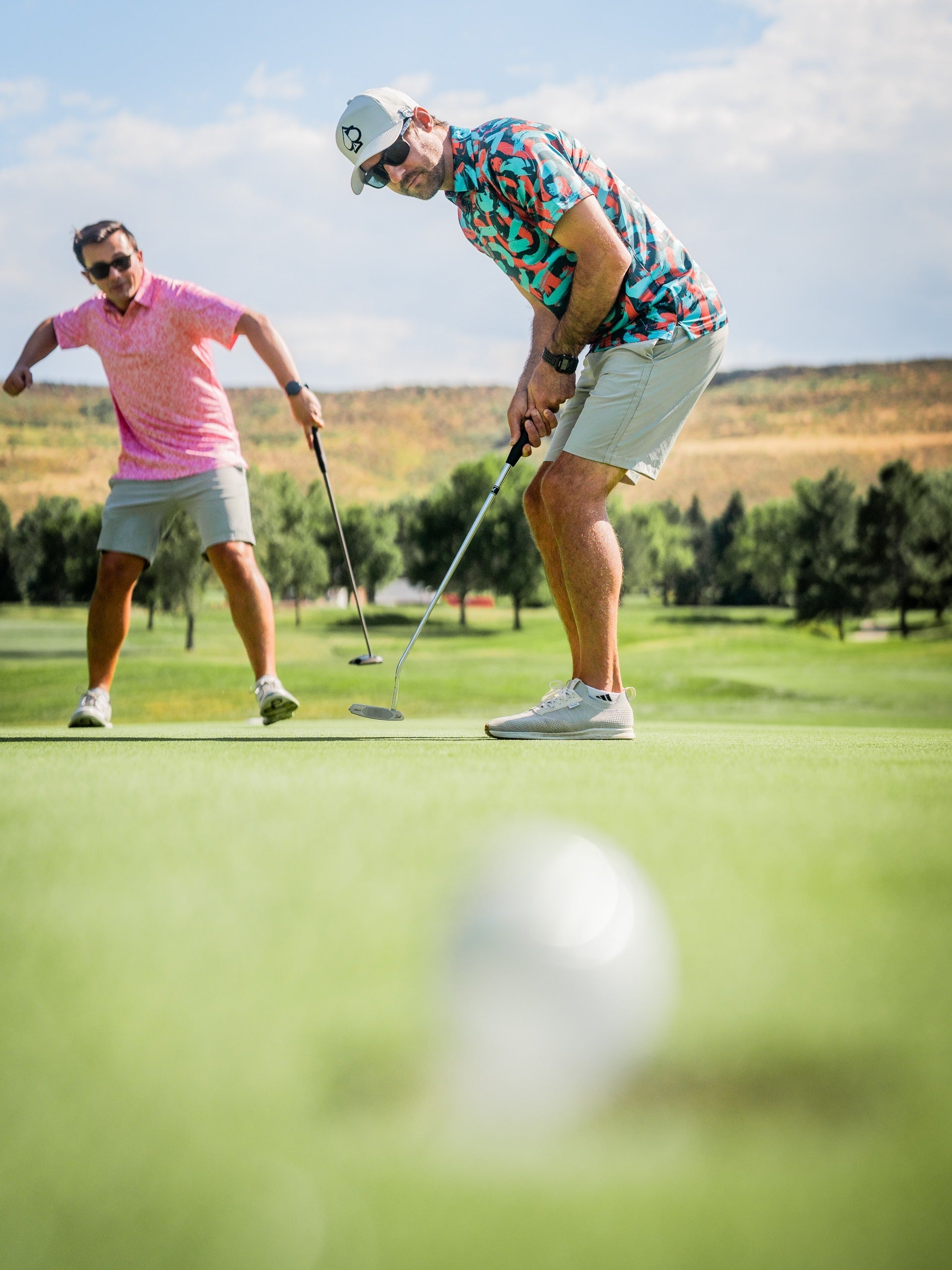 Two golfers on a green, one wearing a colorful Flower Doodle Pink shirt, focusing on a putt while the other watches.