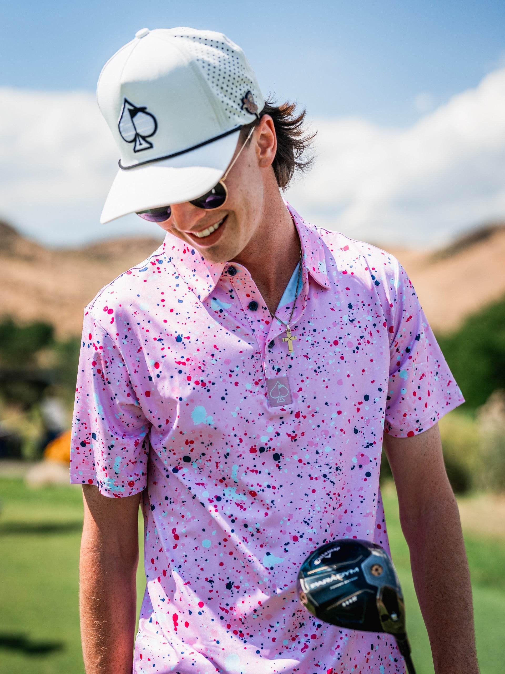 A golfer wearing a pink splatter polo shirt and a cap smiles while holding a golf club on a sunny course.
