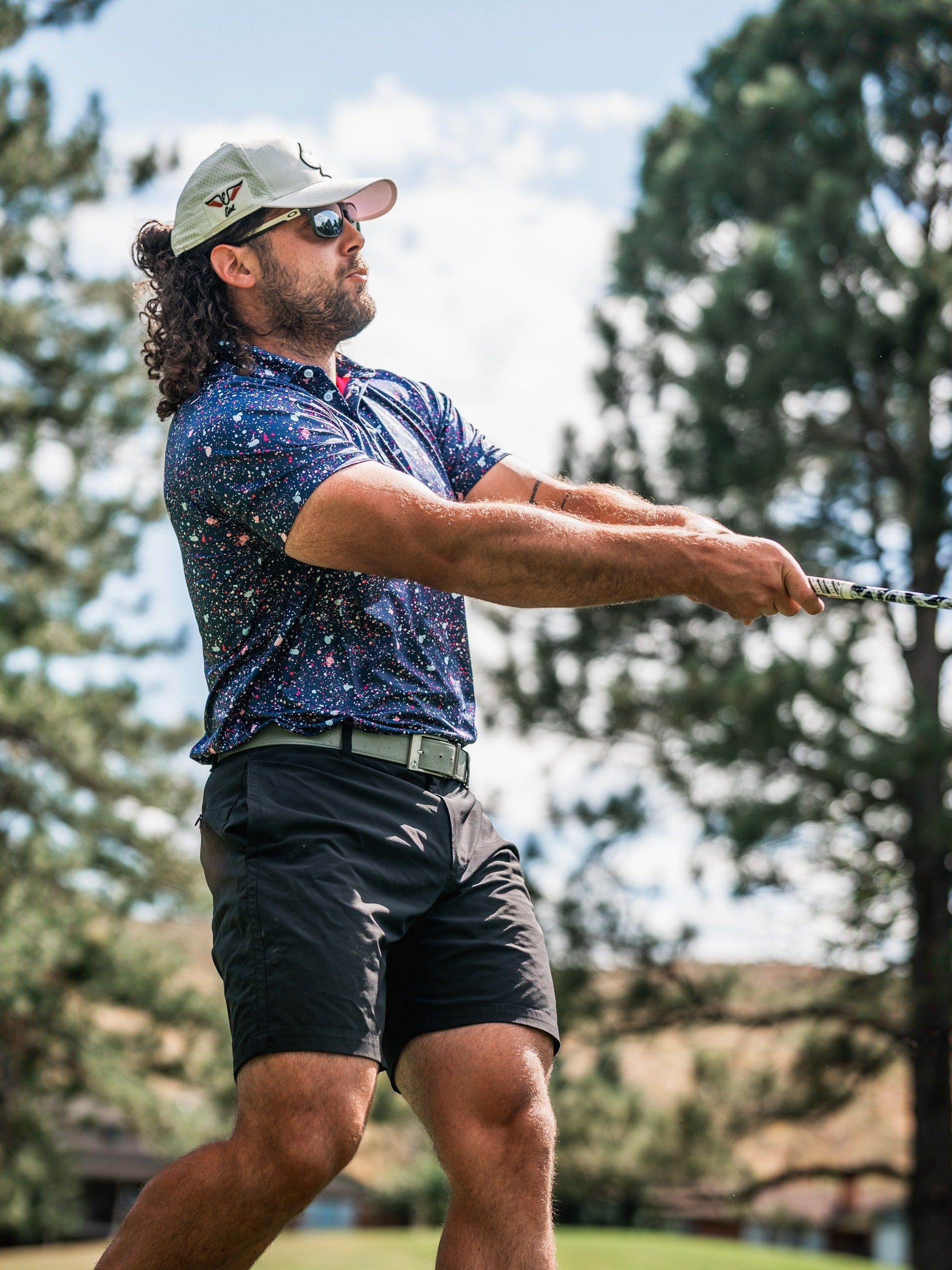 A golfer swings a club wearing a blue splatter-patterned polo shirt and shorts, surrounded by trees and a clear sky.
