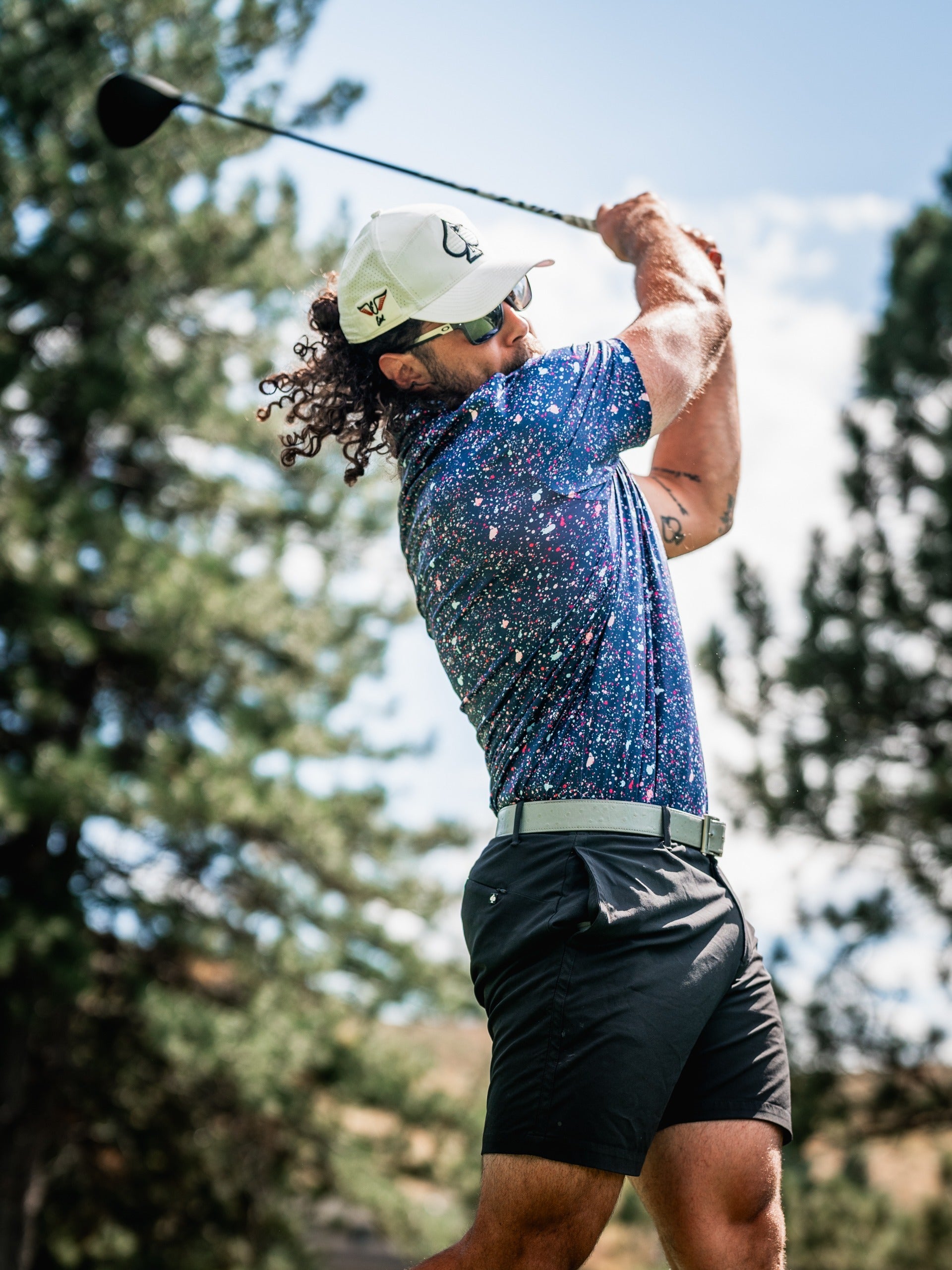 A golfer swings a driver, wearing a blue splatter-patterned polo shirt and shorts, surrounded by trees on a sunny day.