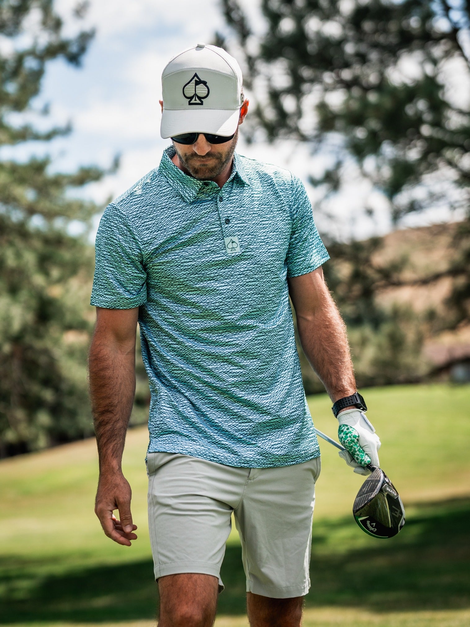Stylish golfer wearing a teal squiggle-patterned polo and light shorts, holding a golf club on a sunny course.