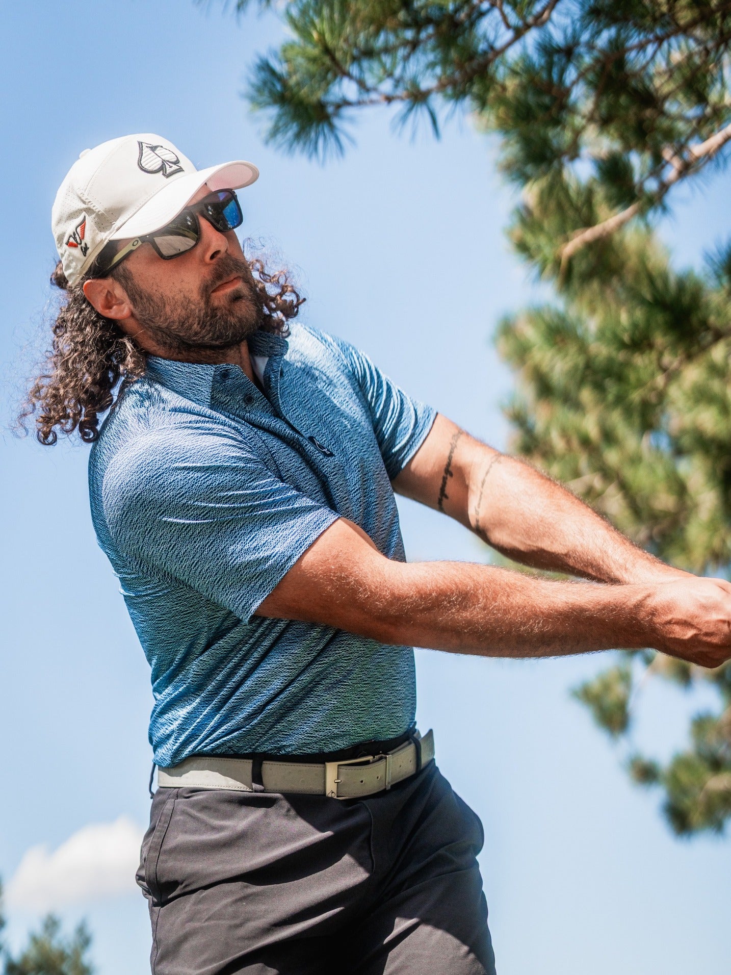 A golfer swings a club while wearing a blue patterned polo shirt and a cap, showcasing a stylish look on the course.