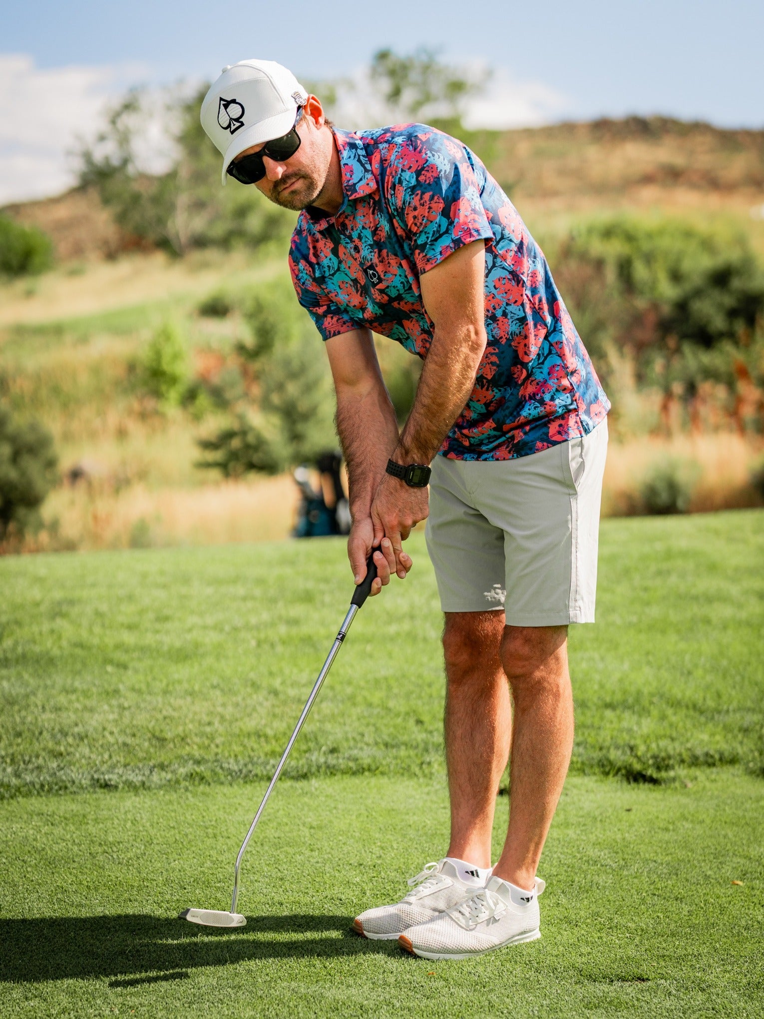 Man in colorful floral golf shirt, white shorts, white sneakers, white cap with black logo, and sunglasses on a golf course.