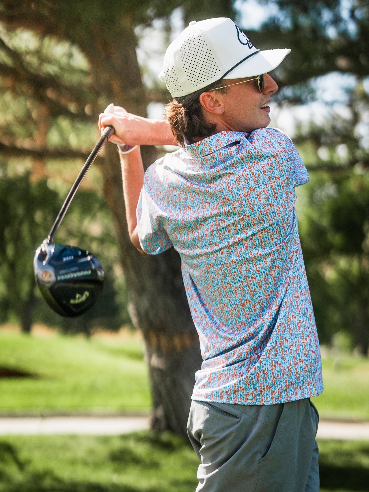 Male golfer in colorful patterned shirt and white cap swings club on a golf course with trees in background.