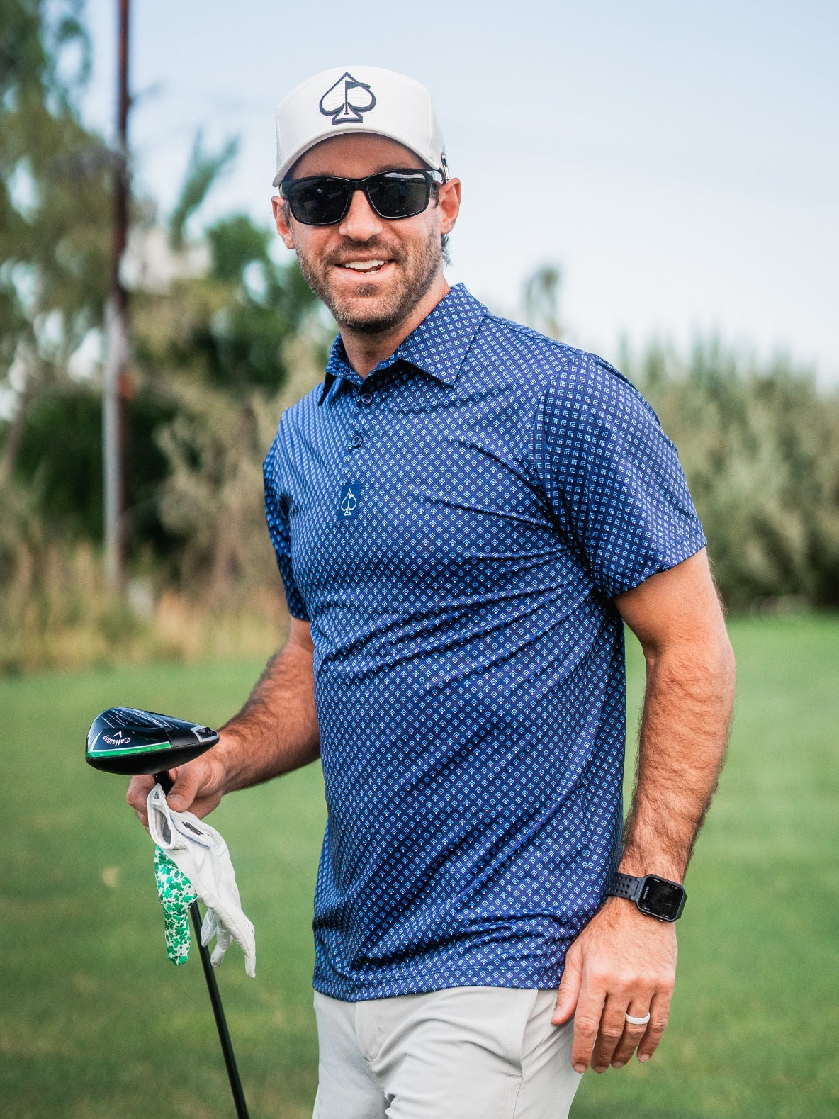 Man wearing a blue patterned golf polo, white cap with spade logo, black sunglasses, and black watch, holding a golf club ...