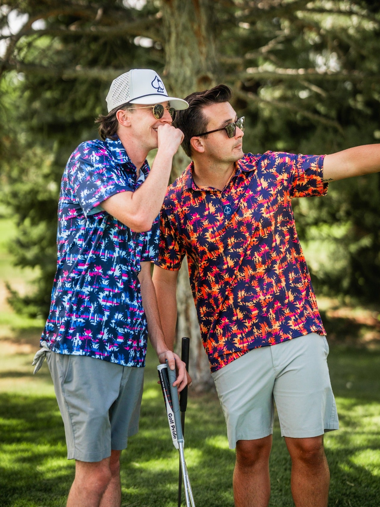 Two men in colorful, tropical-patterned golf shirts, wearing sunglasses, outdoors on a sunny day, discussing or pointing a...
