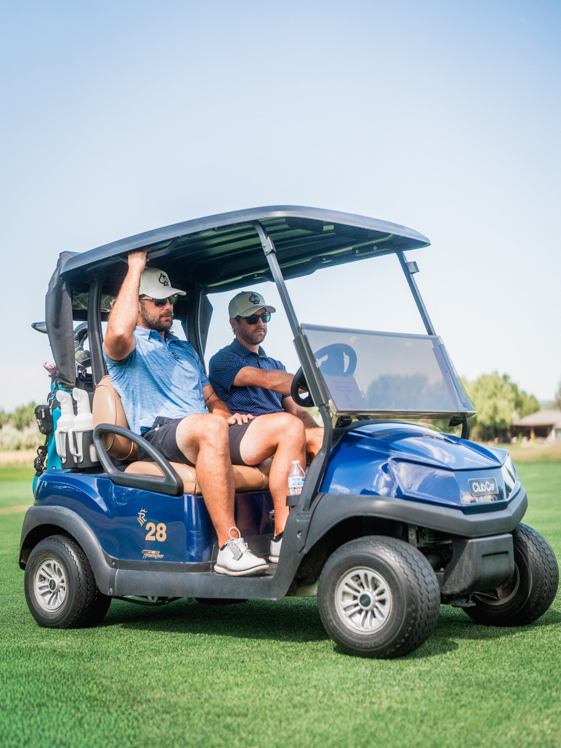 Two men in golf attire sit in a blue golf cart on a lush green course, enjoying a sunny day.