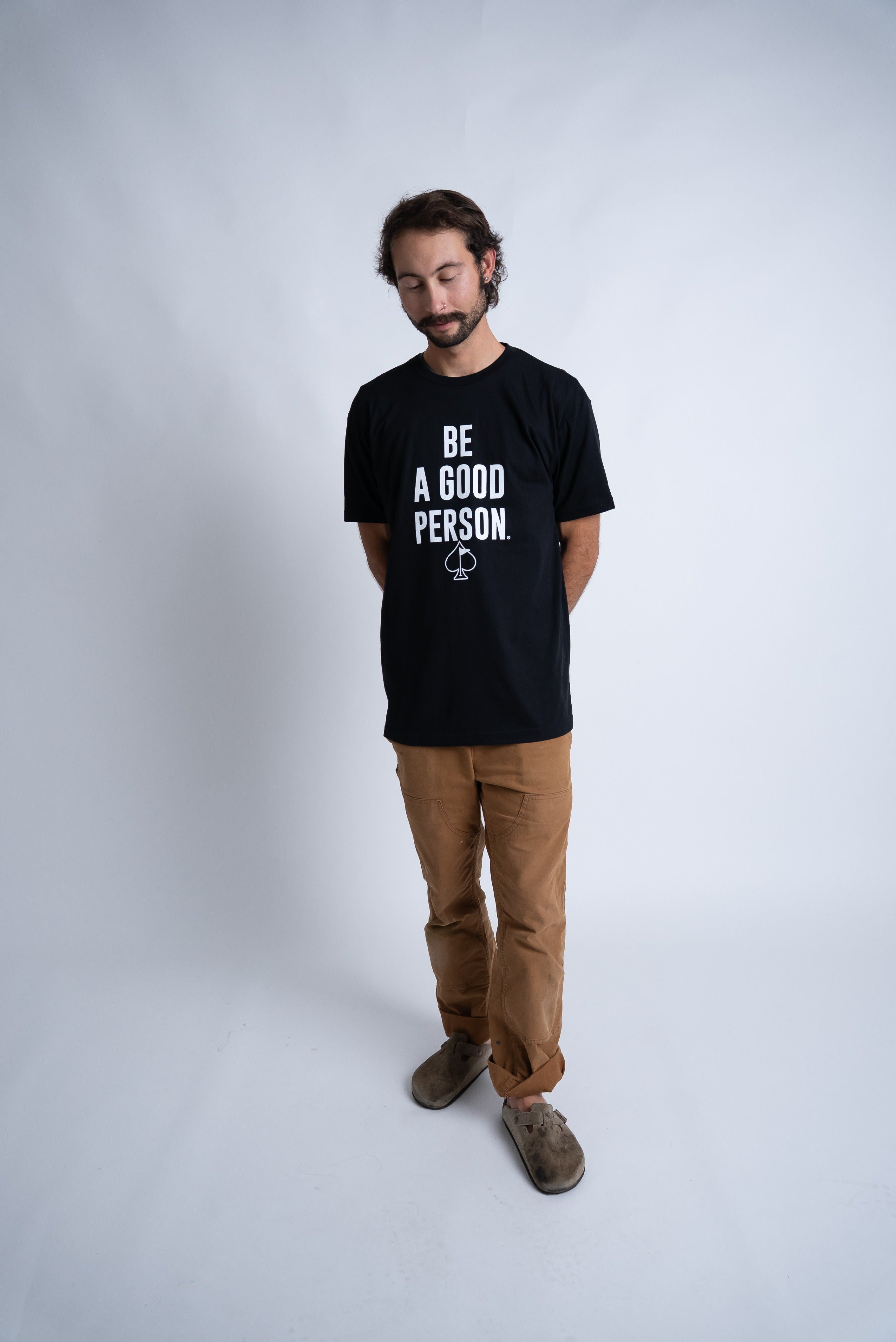 Black t-shirt featuring the phrase "Be A Good Person" in bold white letters, worn by a man standing casually.