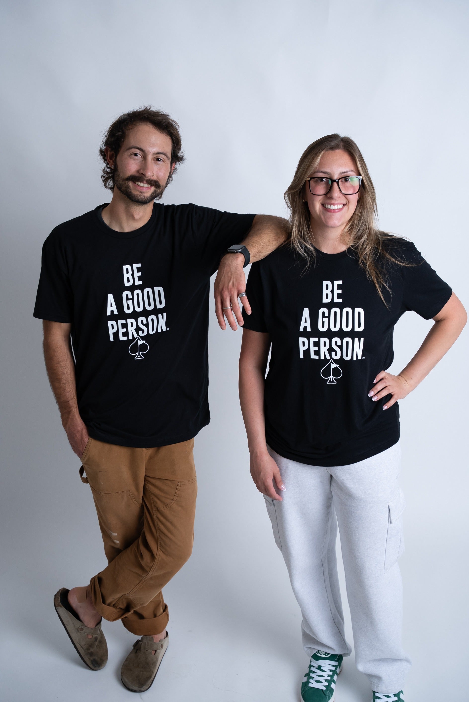 Two people wearing matching black t-shirts that read "BE A GOOD PERSON" in bold white letters, posing together.