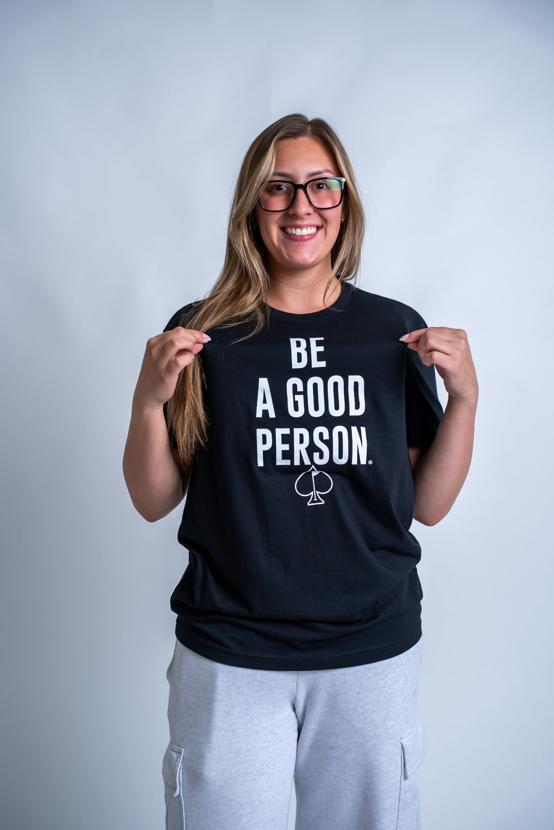 Black t-shirt featuring the phrase "Be A Good Person" in bold white letters, worn by a smiling person.