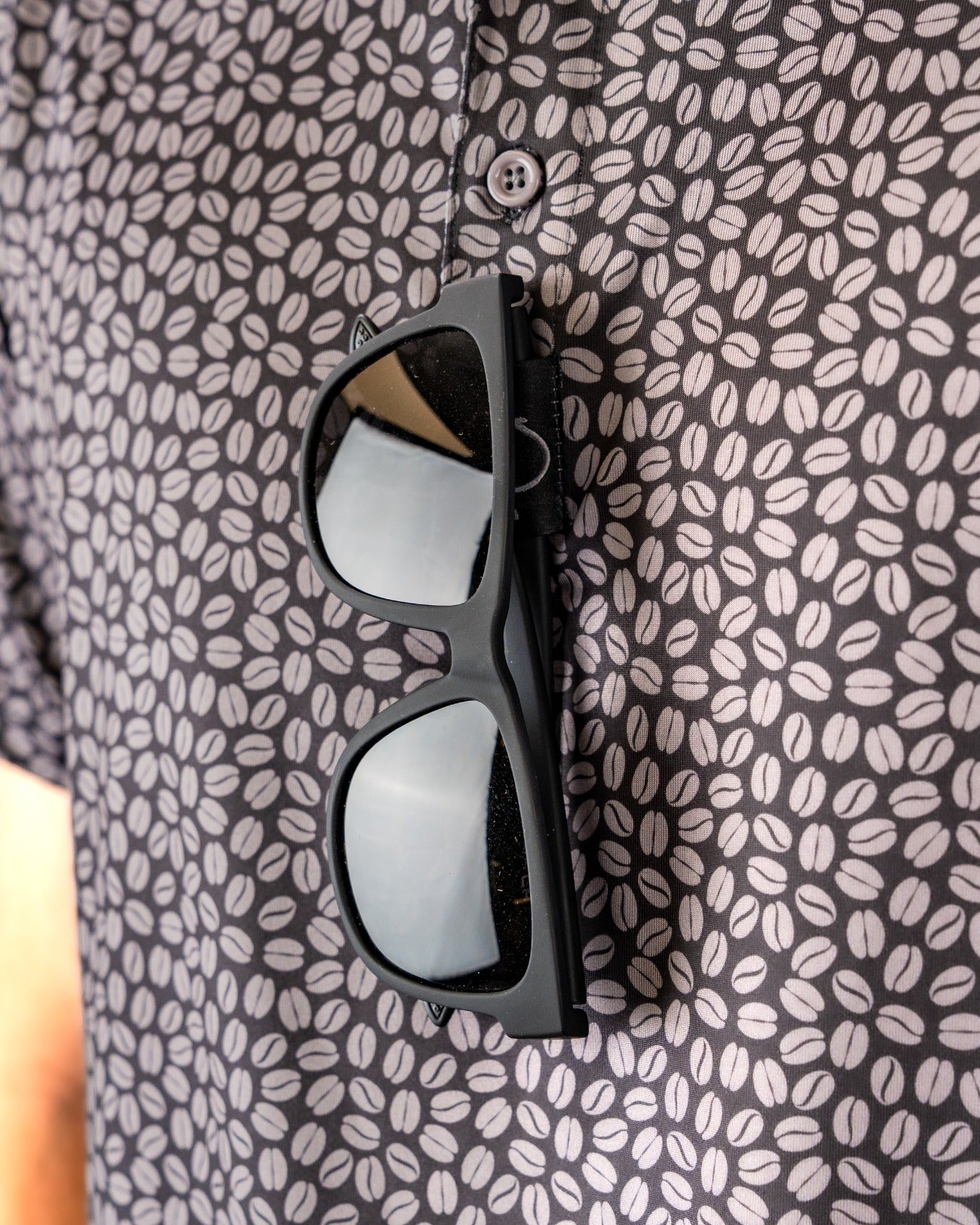 Black polo shirt with a coffee bean pattern, featuring sunglasses resting on the collar.