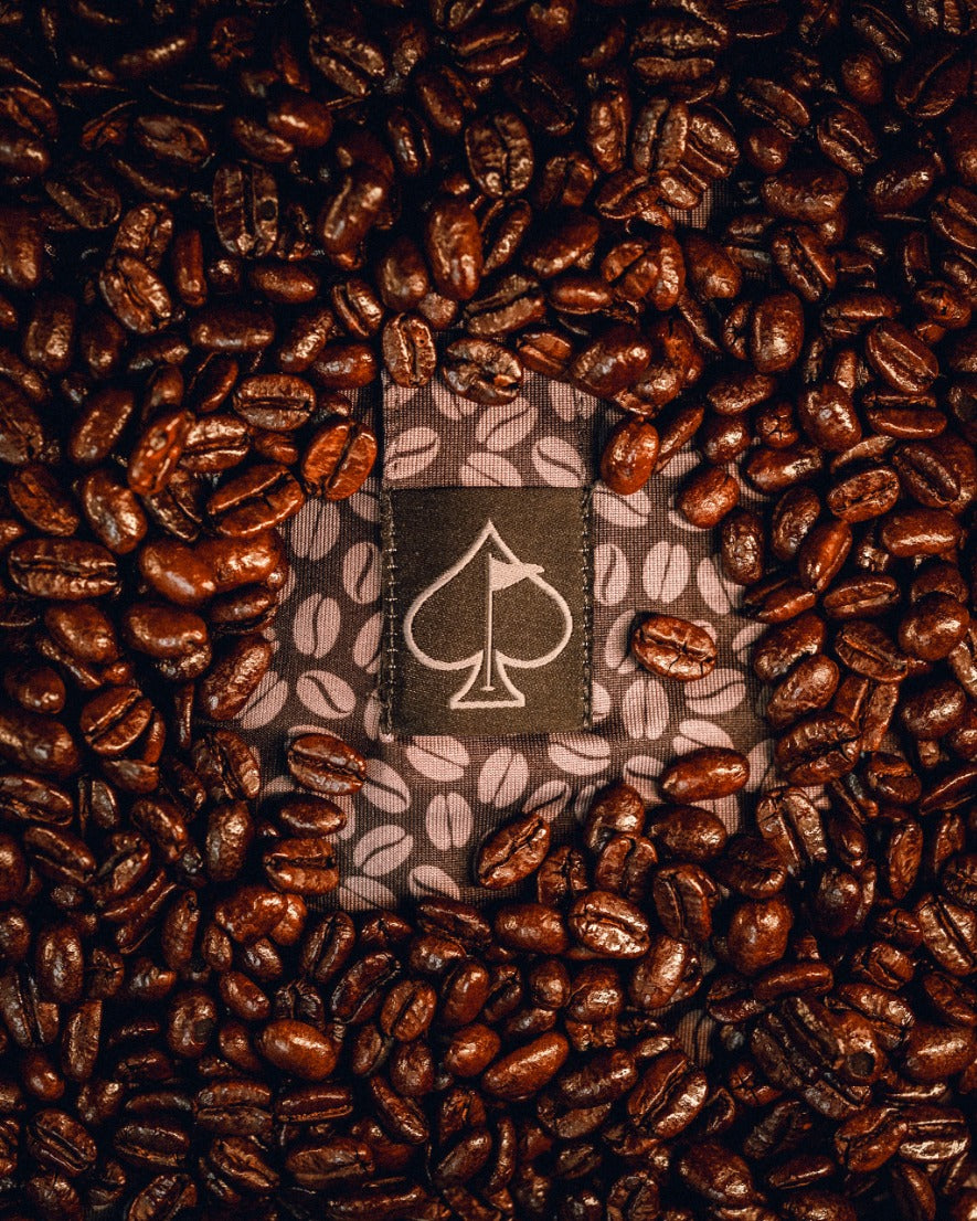 A Generous Coffee polo shirt surrounded by dark roasted coffee beans, featuring a logo of a spade.