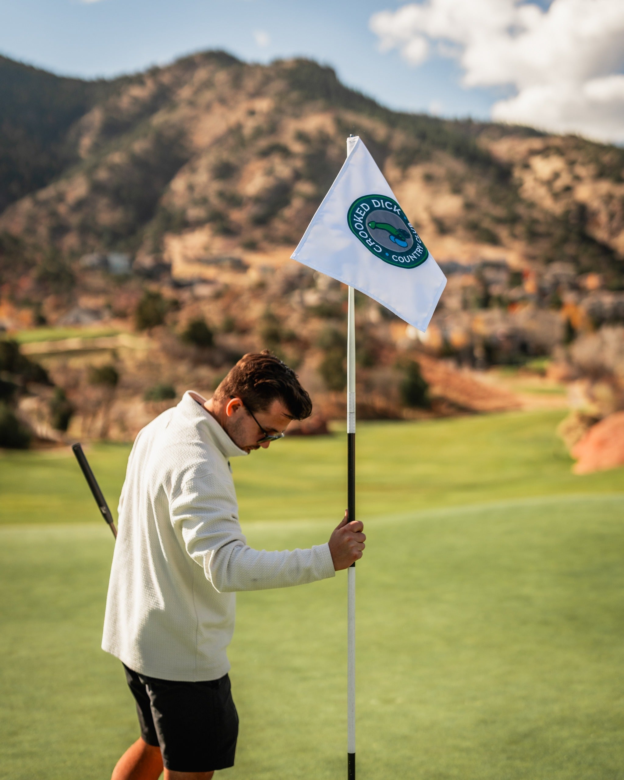 A golfer walks on the green, holding a Crooked Dick golf flag with a scenic mountain backdrop.