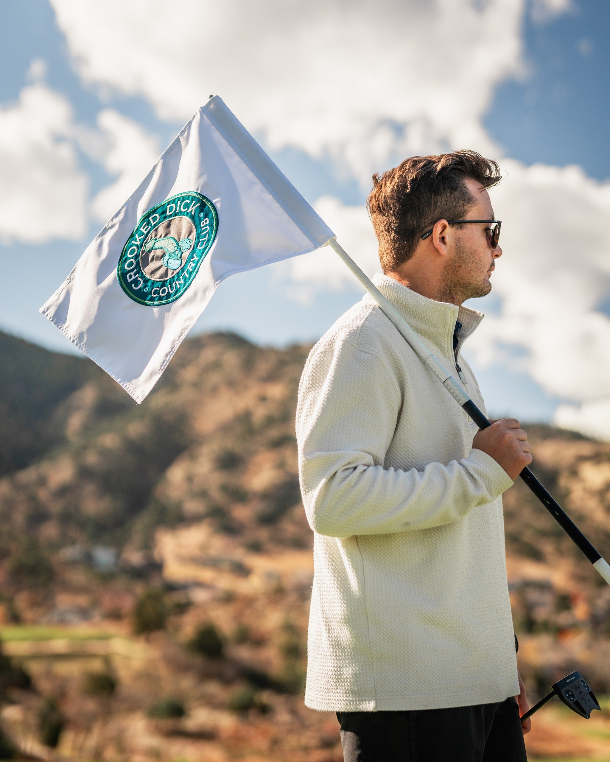 A golfer stands on the course holding a Crooked Dick golf flag, showcasing its unique design against a scenic backdrop.