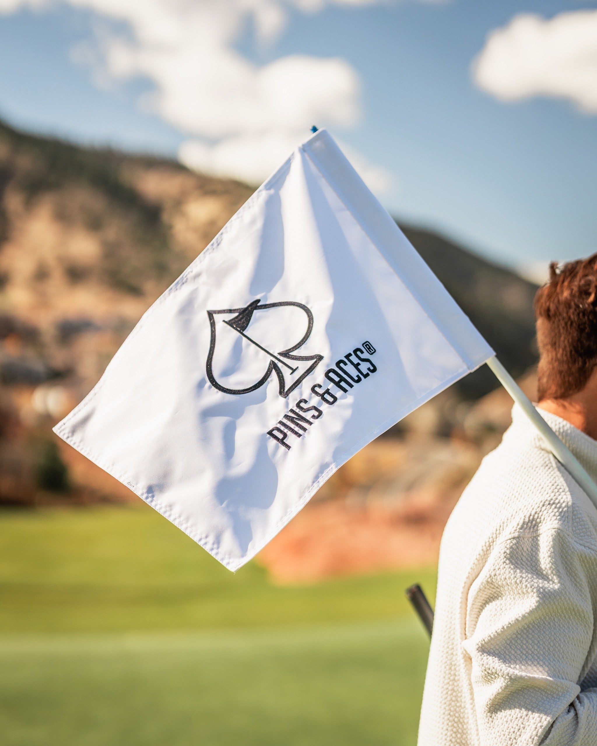White golf flag featuring the Pins and Aces logo, held by a golfer on a sunny course with mountains in the background.
