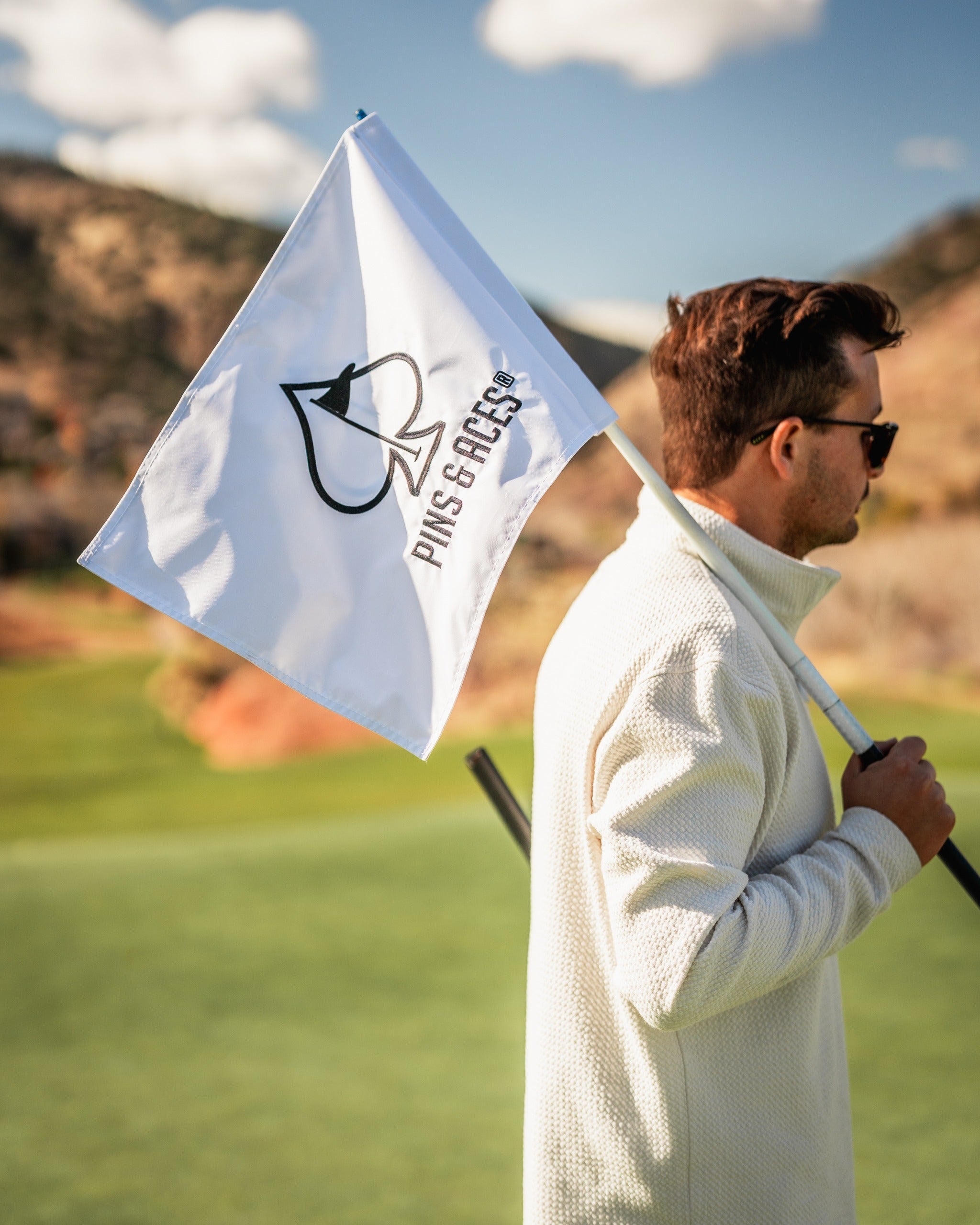 A golfer holds a Pins and Aces golf flag on the course, showcasing the brand's logo against a scenic backdrop.