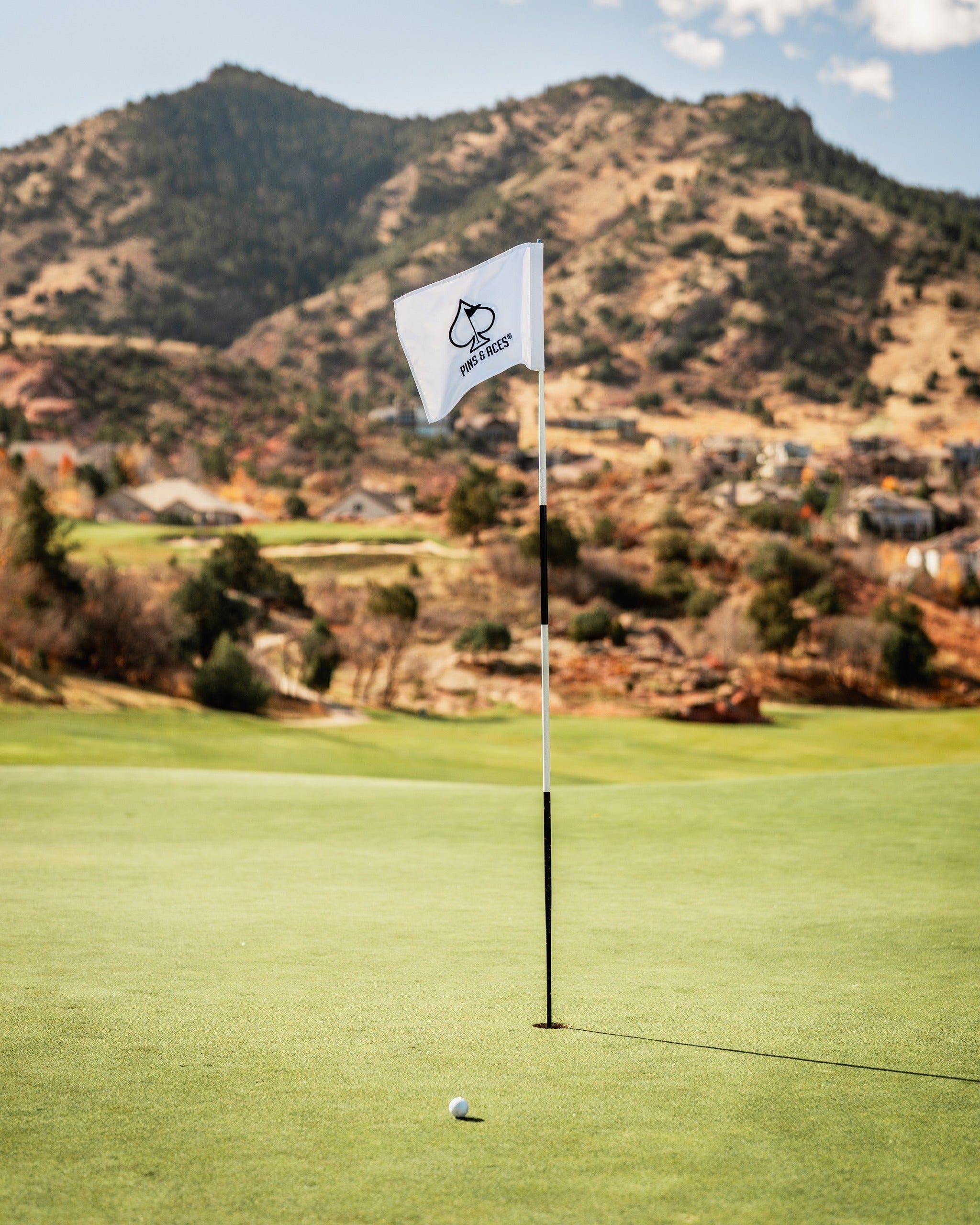 Golf flag with Pins and Aces logo on a lush green course, set against a backdrop of mountains and blue sky.