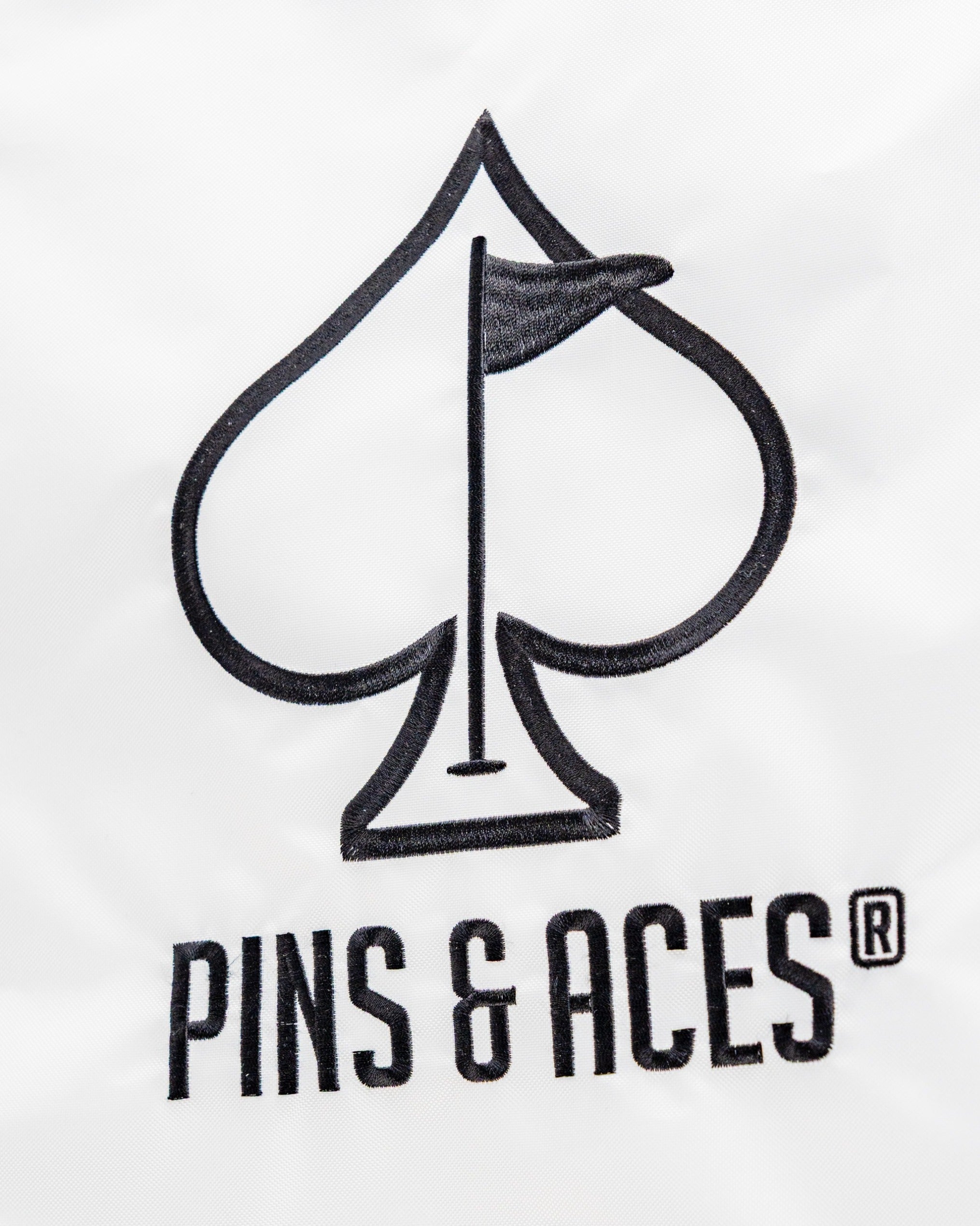 Embroidered Pins and Aces logo featuring a golf flag inside a spade symbol on a white background.