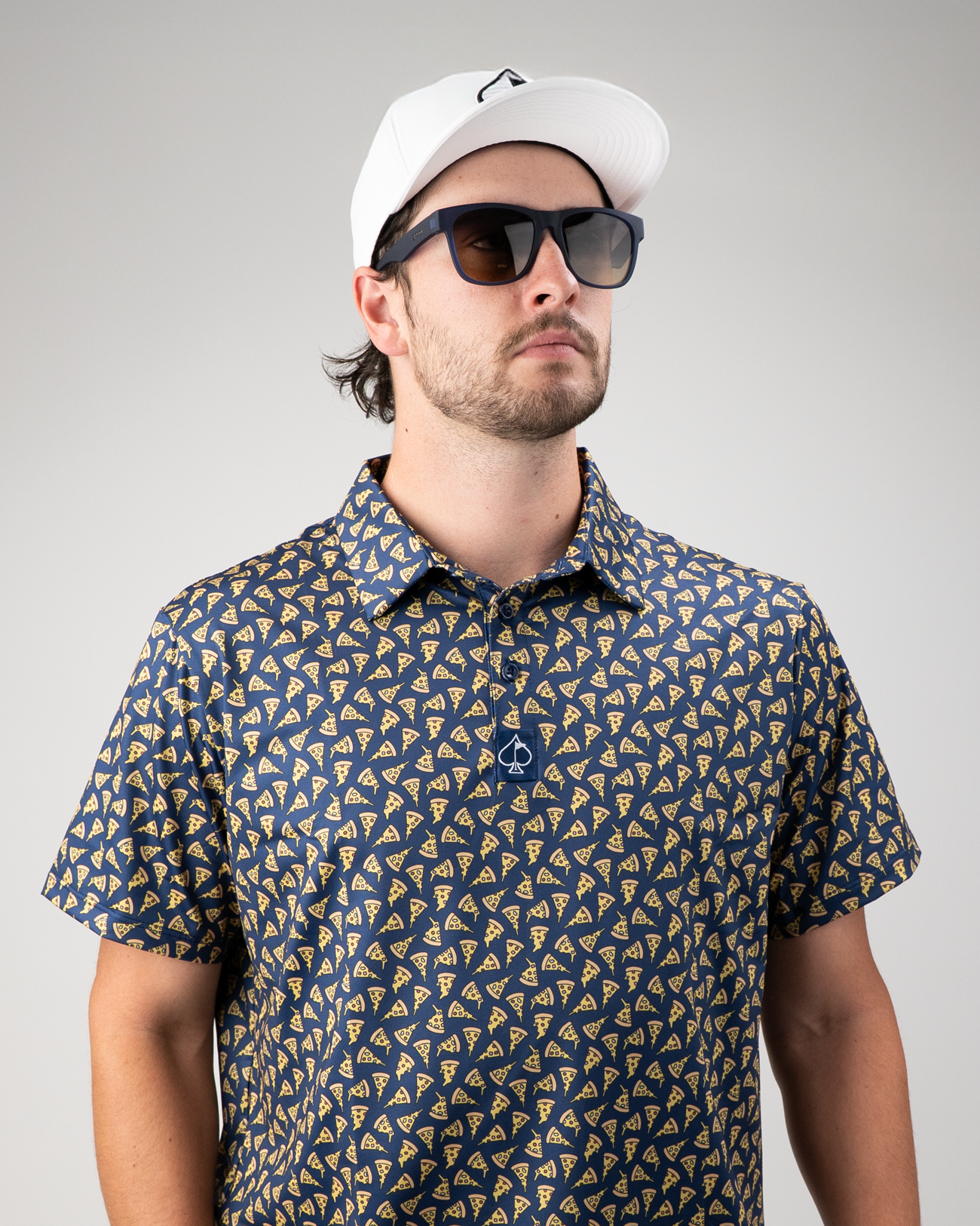 Man wearing a navy golf polo with a pizza slice pattern, sunglasses, and a white cap, posing confidently.