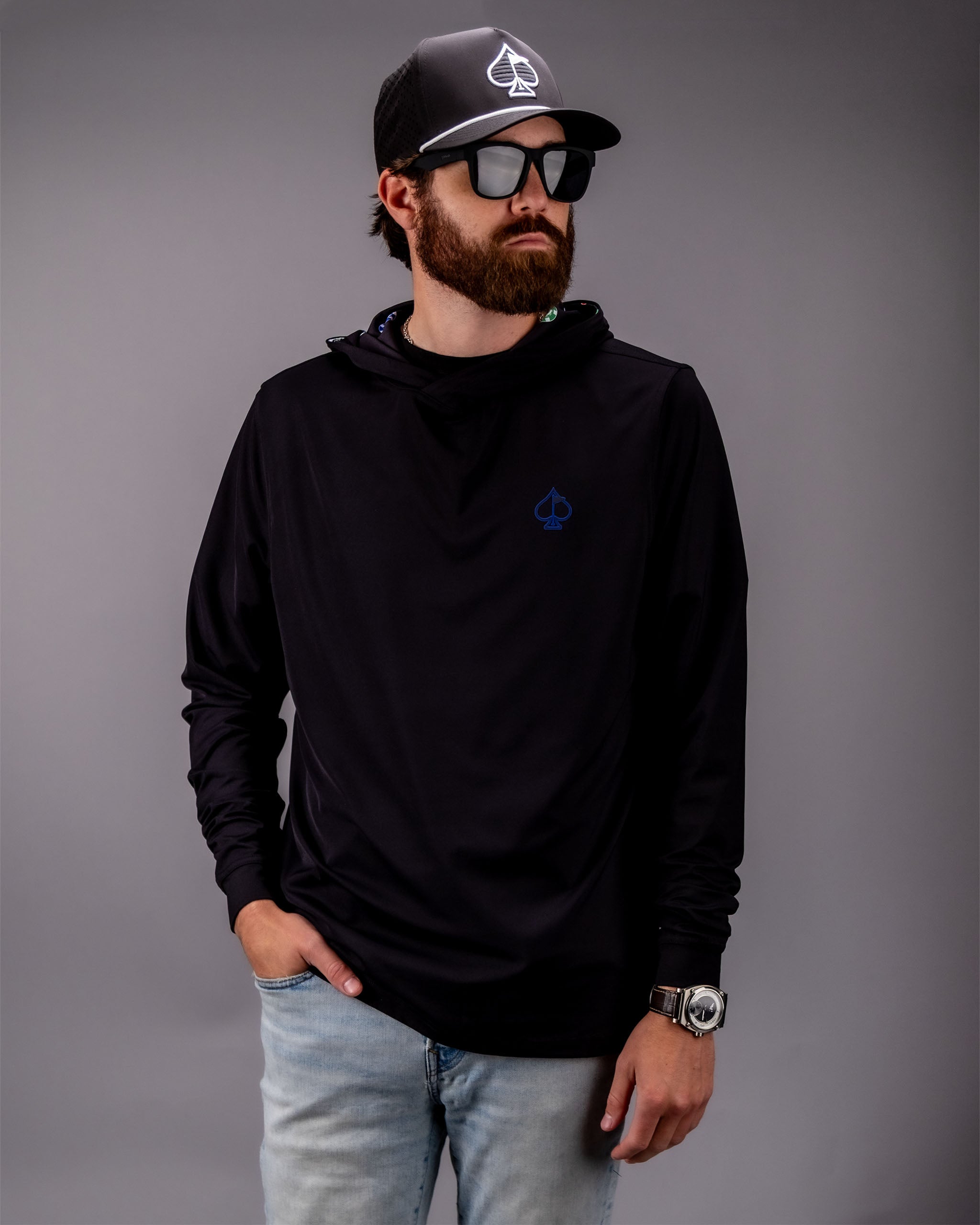 Male model wearing a black performance golf hoodie with a blue spade logo, paired with a black cap, sunglasses, and light jeans.