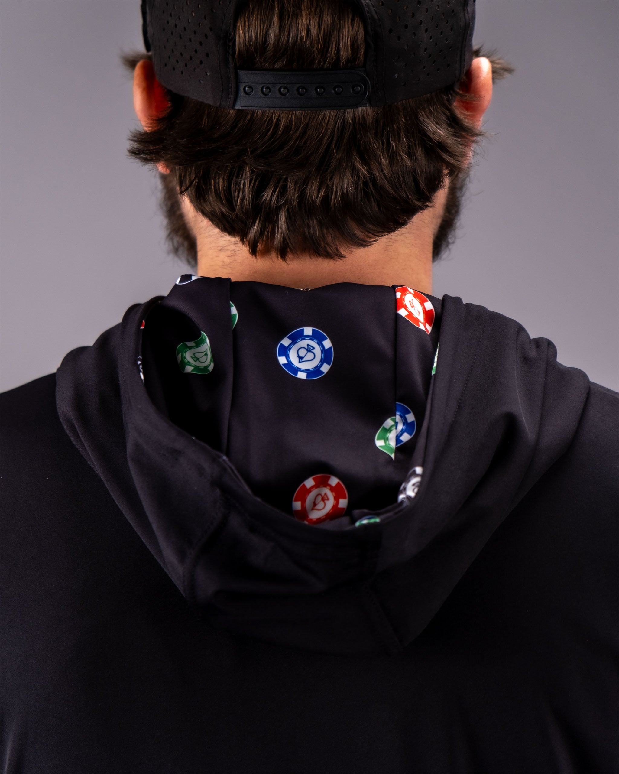 Back view of a person wearing a black performance golf hoodie with poker chip patterns and a black cap.