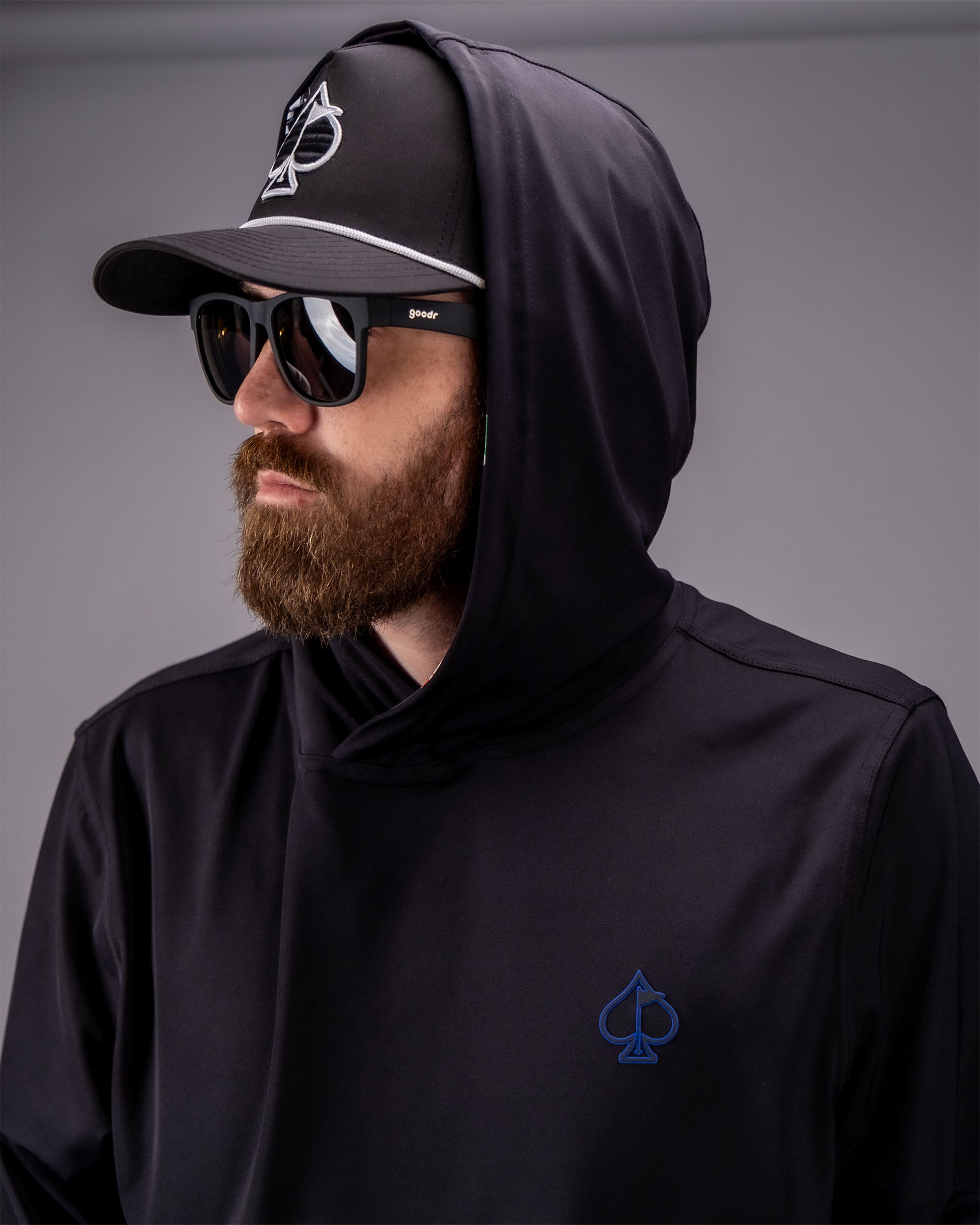 Man wearing a black performance golf hoodie with a blue spade logo, black cap, sunglasses, and a beard.