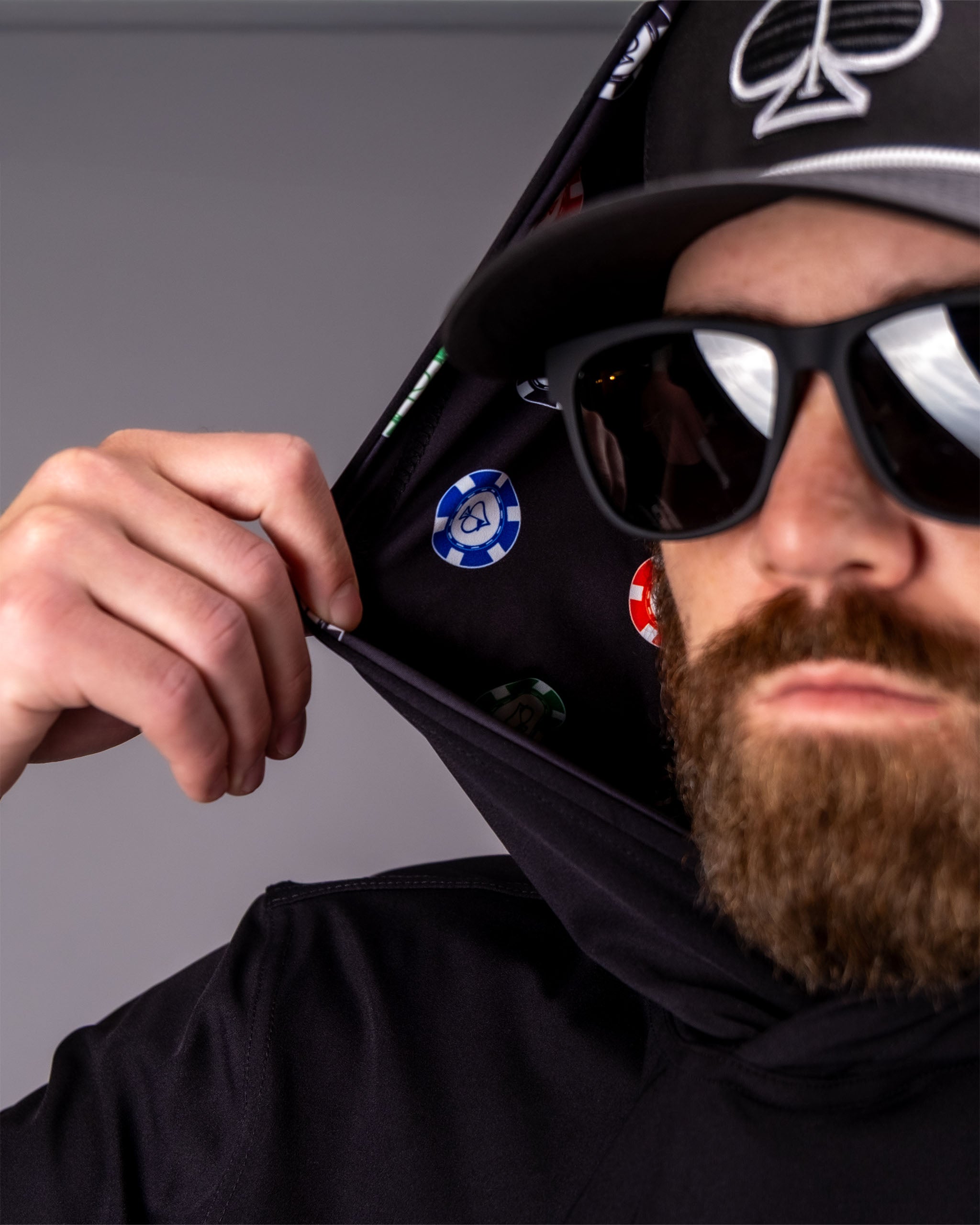 Man wearing a black performance golf hoodie with poker chip designs, sunglasses, and a cap, pulling the hoodie away from his face.