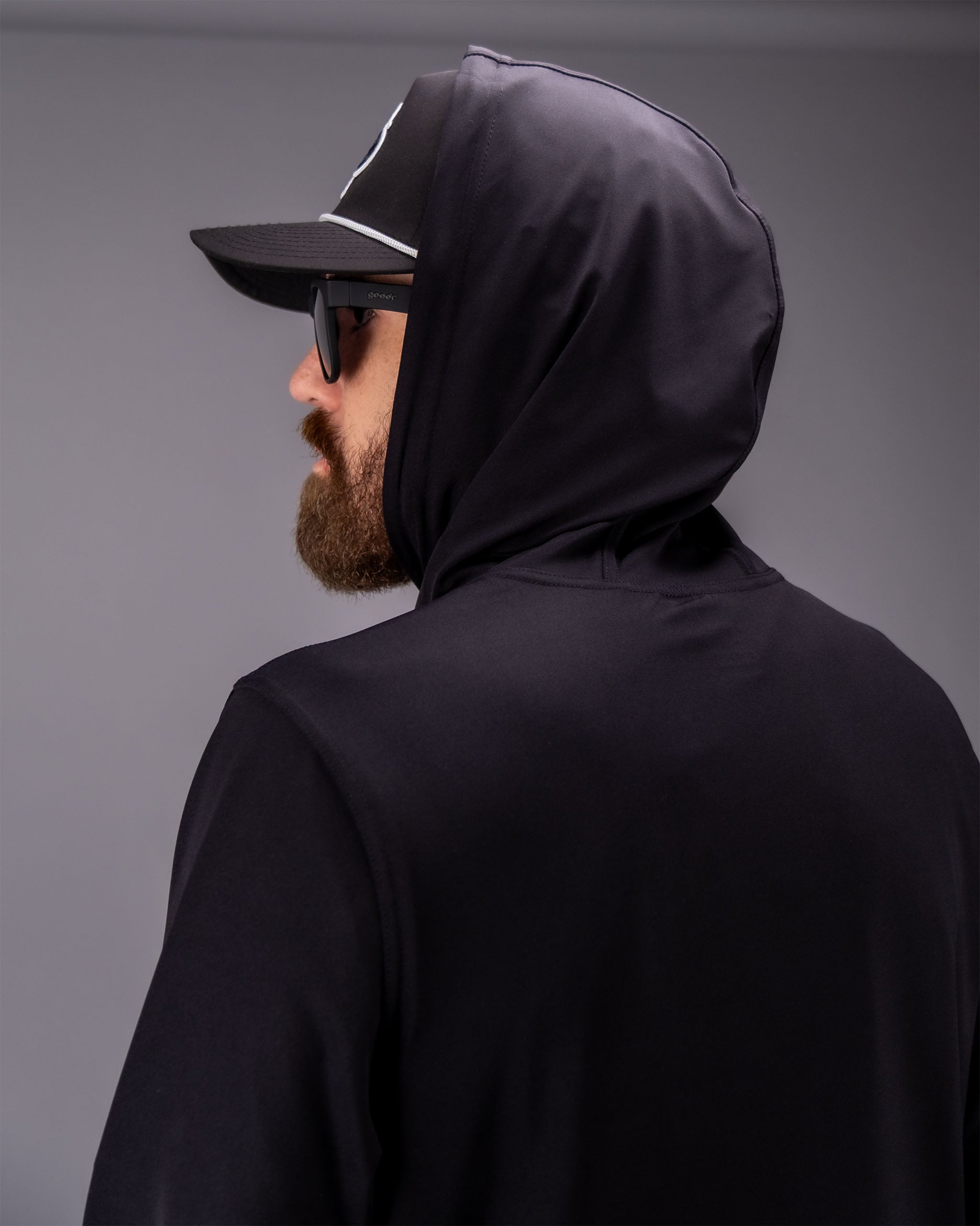 Man wearing a black performance golf hoodie with a hood, sunglasses, and a cap, shown in profile against a gray background.