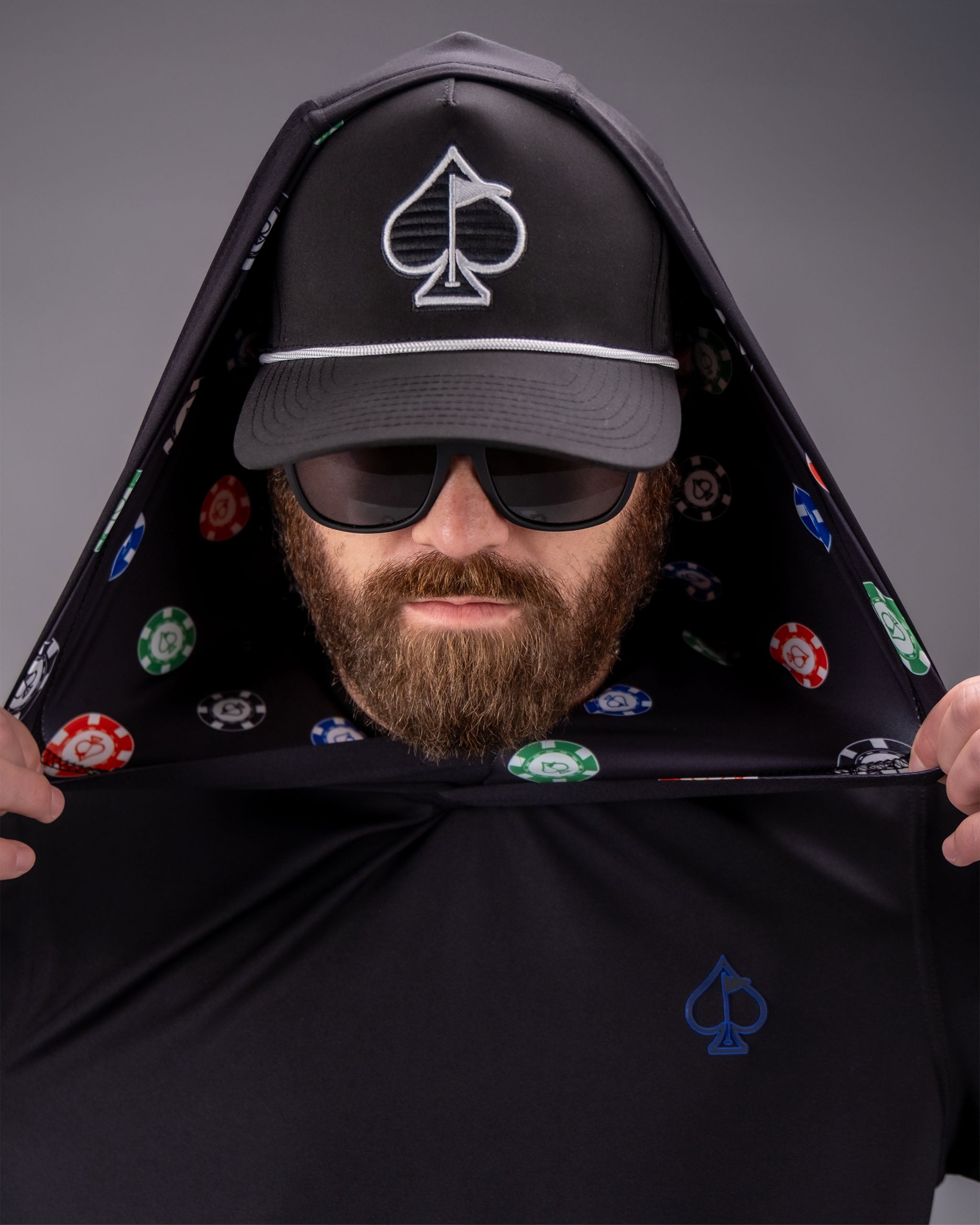Man wearing a black performance golf hoodie with poker chip pattern lining, black cap with spade logo, and sunglasses.