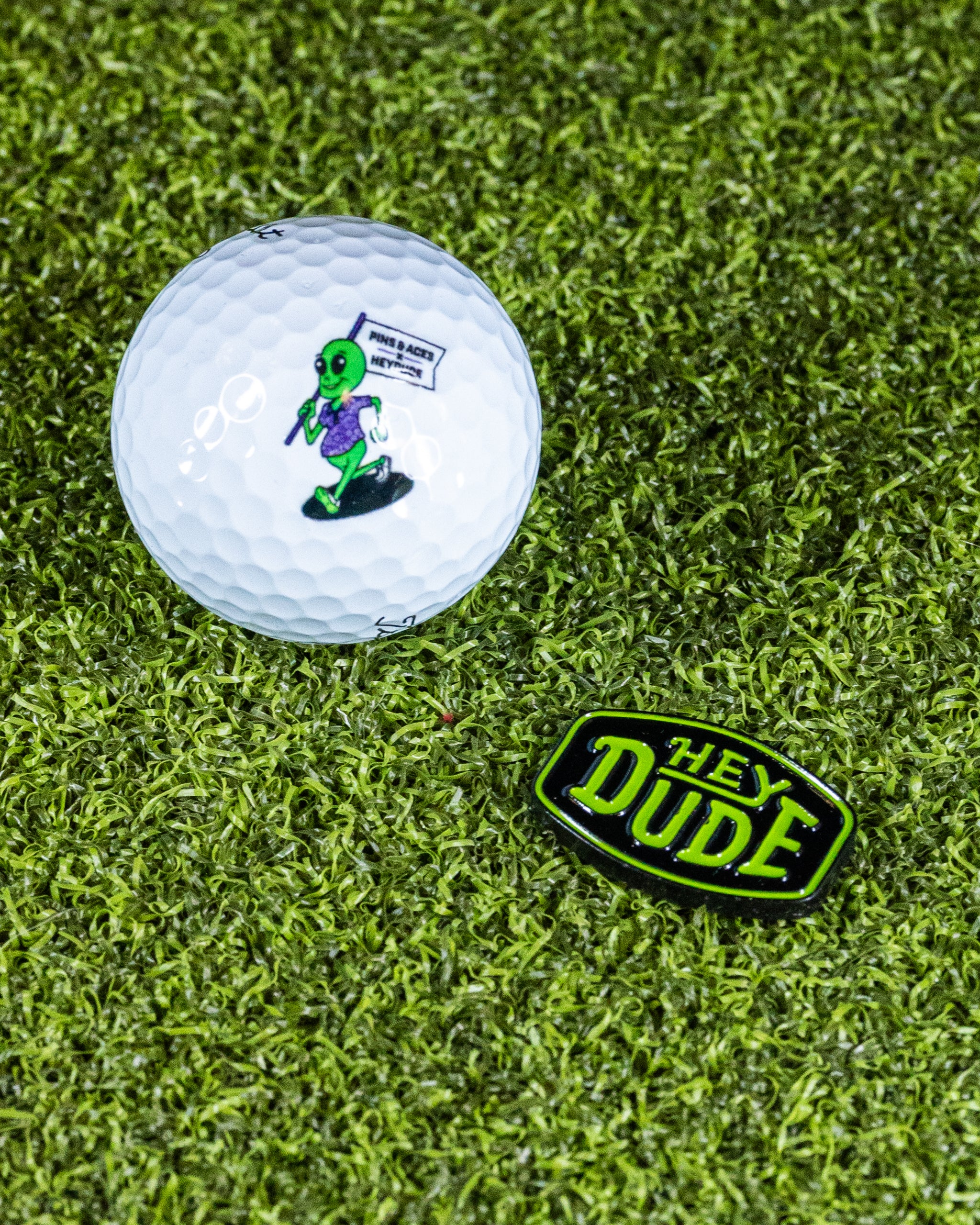 "HEYDUDE logo ball marker on a green background, featuring a circular design with a bold logo."