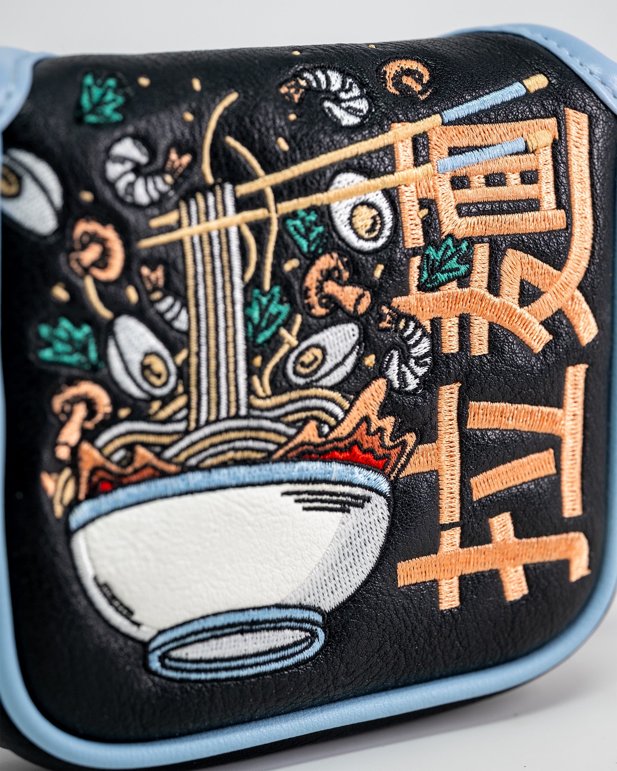 Embroidered patch of a ramen bowl with noodles, chopsticks, and vegetables on a black golf mallet putter cover with light blue trim.