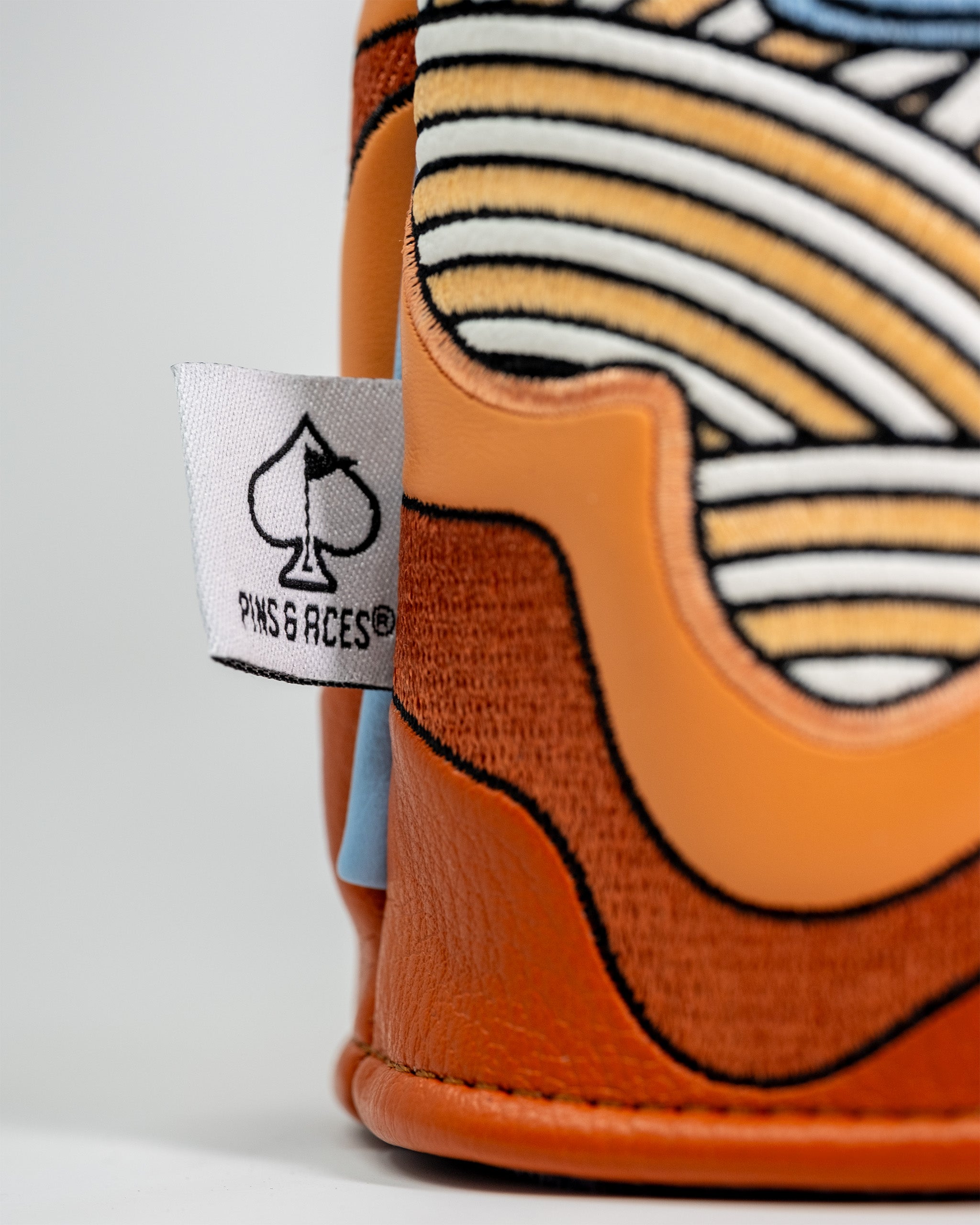 Close-up of a Pins and Aces driver cover featuring a vibrant orange and striped design, with a white fabric tag displaying the brand's spade logo.