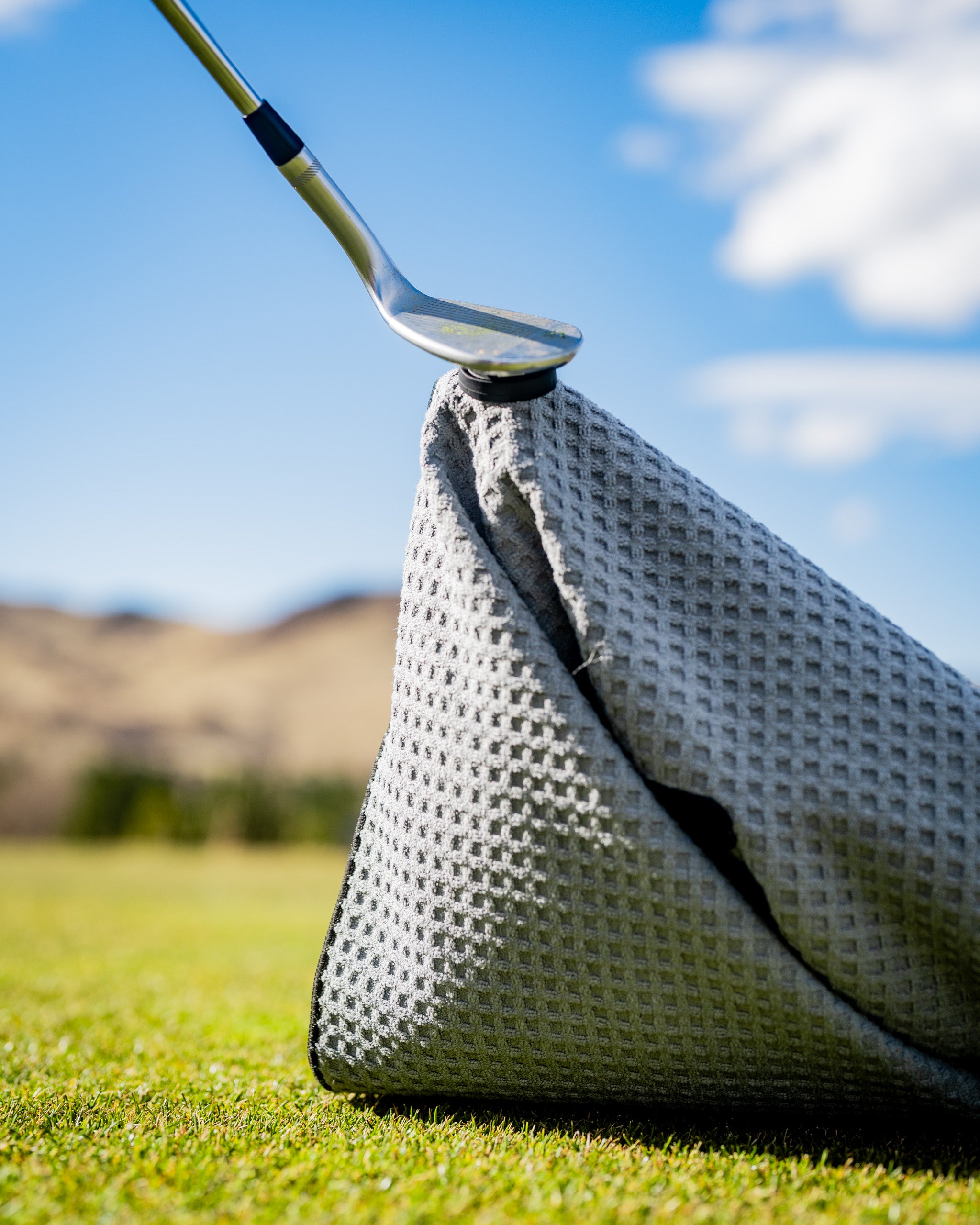 Close-up of a golf club resting on a textured gray headcover on a sunny golf course, with a clear blue sky in the background.