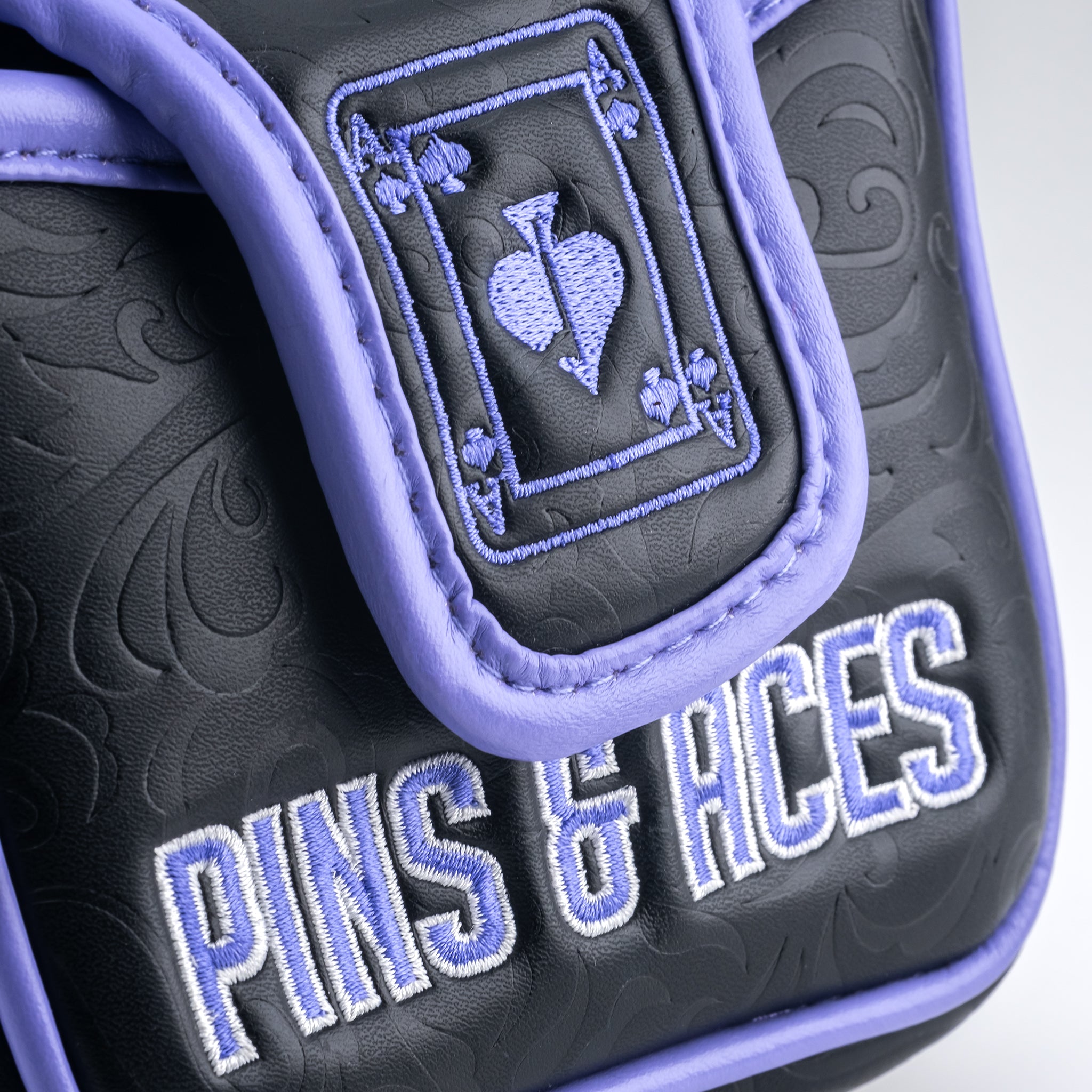 Close-up of a black golf headcover with purple trim, embroidered purple playing card suit symbols, and "PINS & HCES" text.