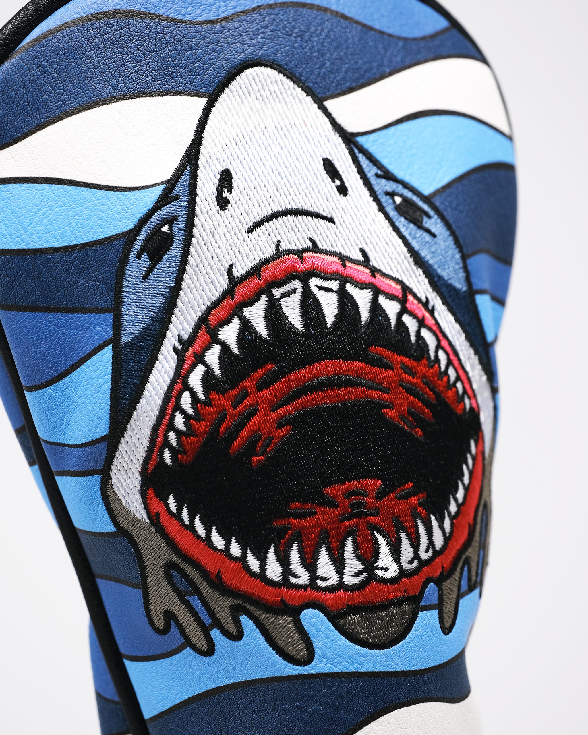 Close-up of a bold, embroidered shark head design on a blue golf accessory, featuring sharp teeth and vivid red, black, an...