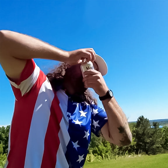 Golf enthusiast wearing a bold American flag-themed shirt, holding a drink, with lush green landscape and clear blue sky.