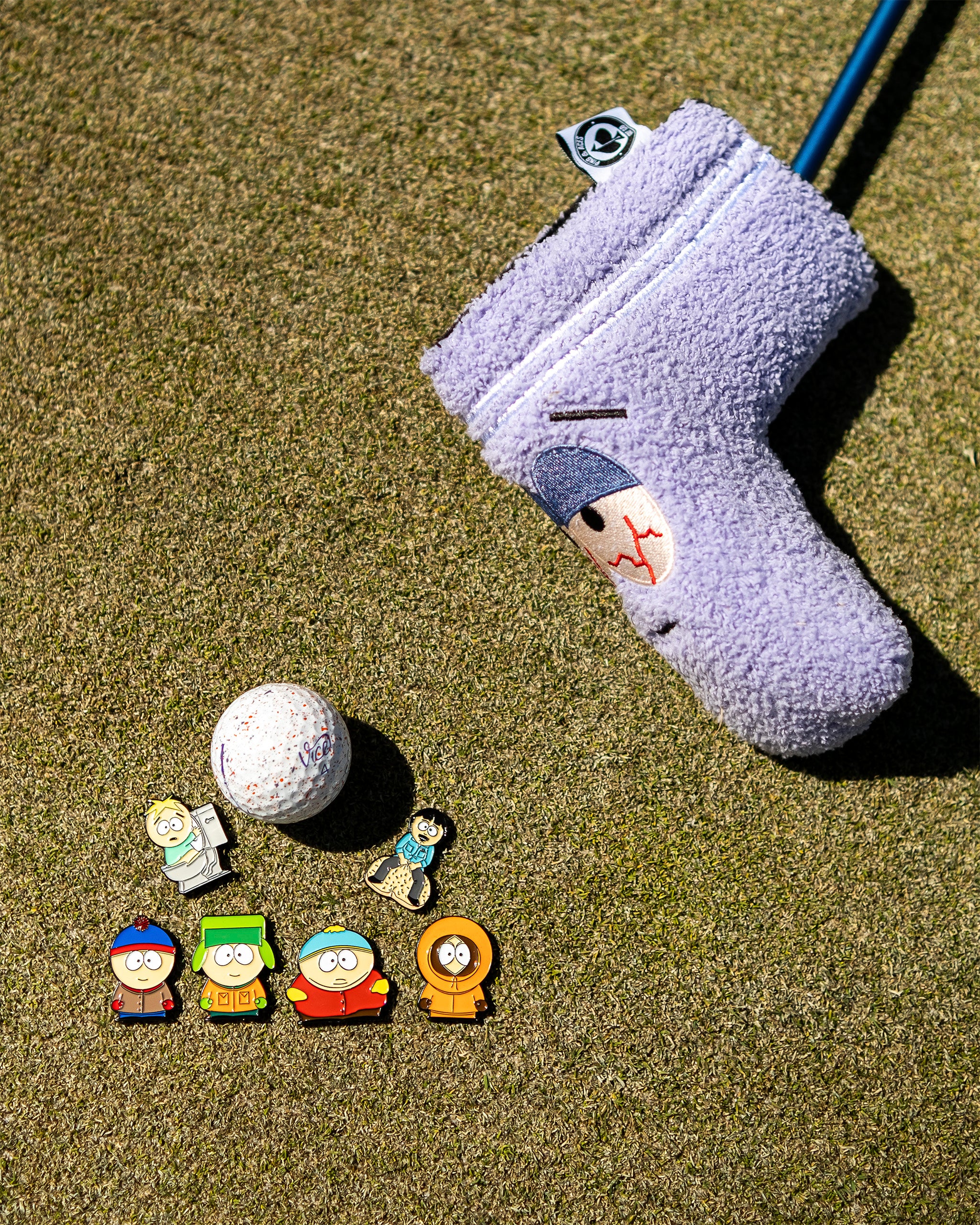 Purple fuzzy golf headcover with a cartoon face, accompanied by golf ball and colorful character pins on a green course.