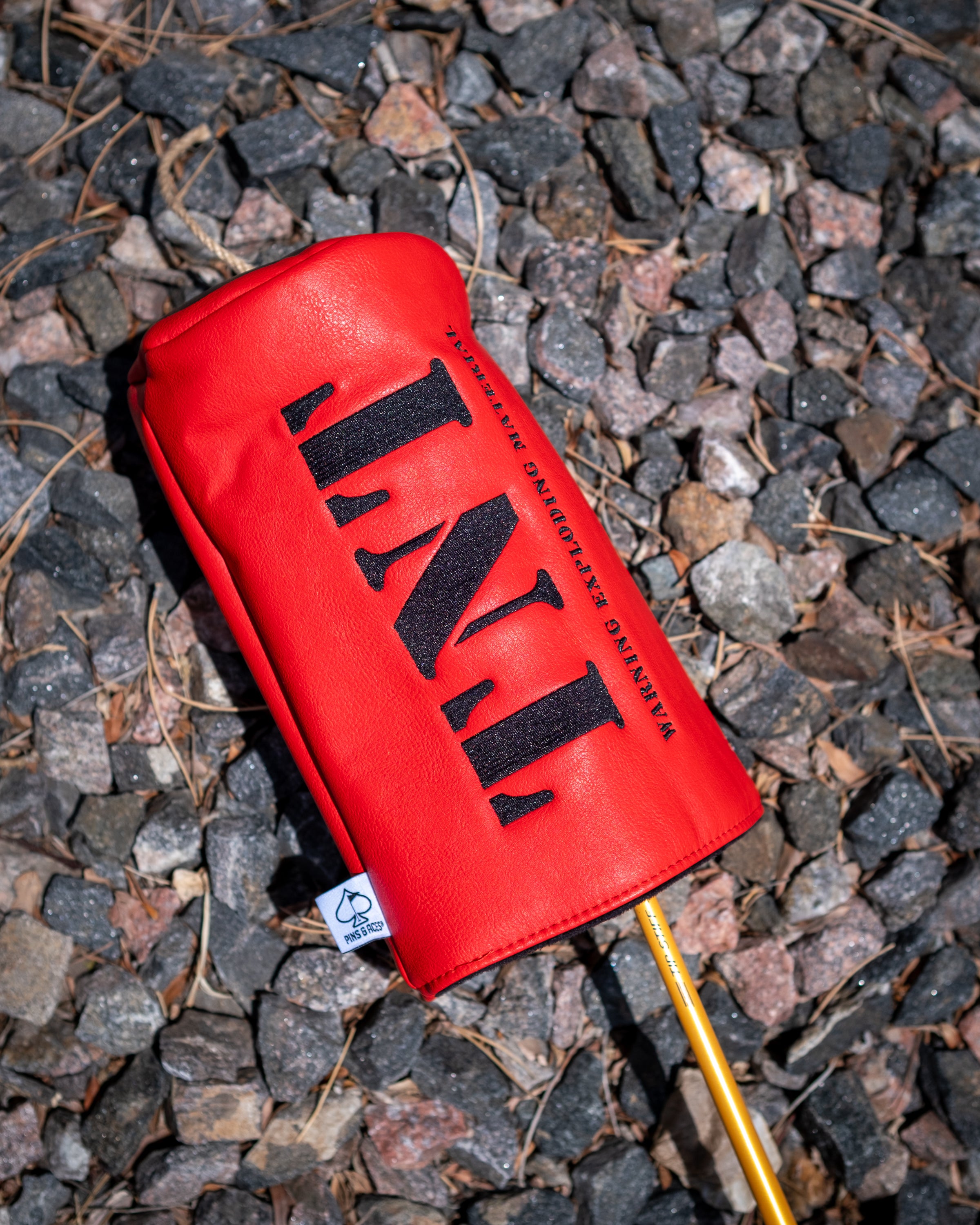 Red driver cover with black "TNT" logo and "Marines Exploding My Life" text, lying on rocky ground with a golf club shaft.
