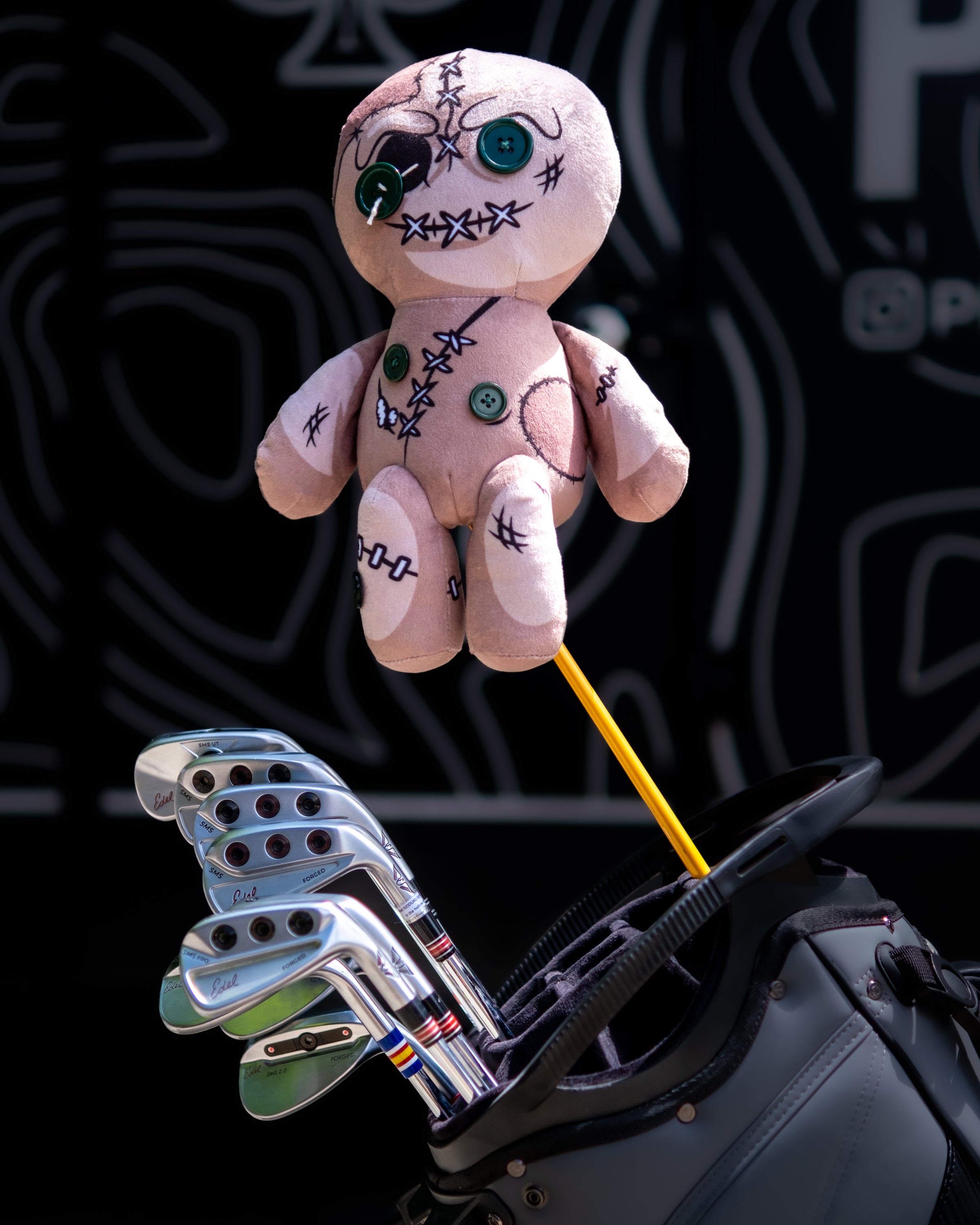 Soft plush voodoo doll driver cover with stitched details and buttons, attached to a golf club bag containing clubs.