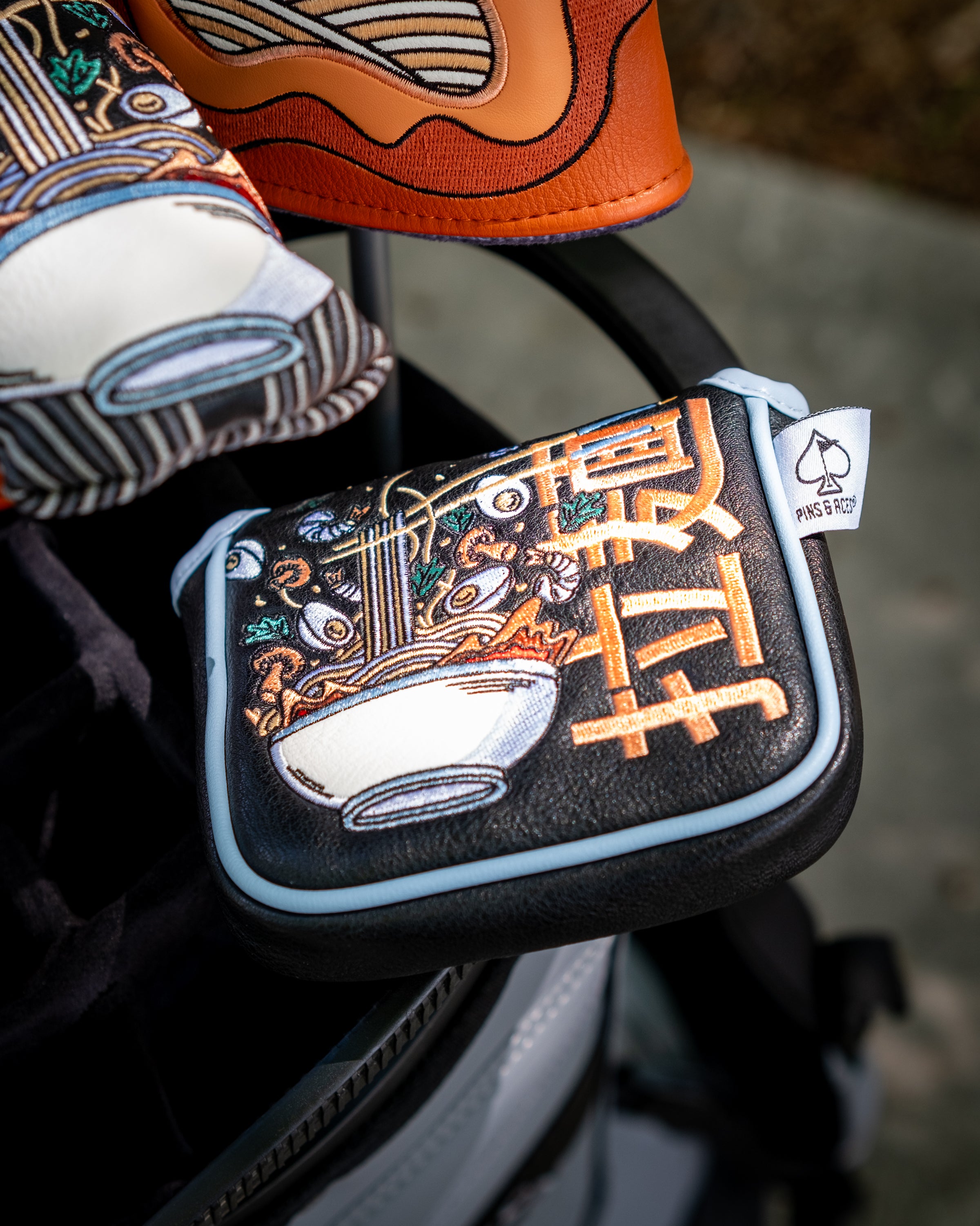 Embossed ramen bowl design on a black mallet putter cover with light blue trim, featuring colorful Asian-inspired embroidery.