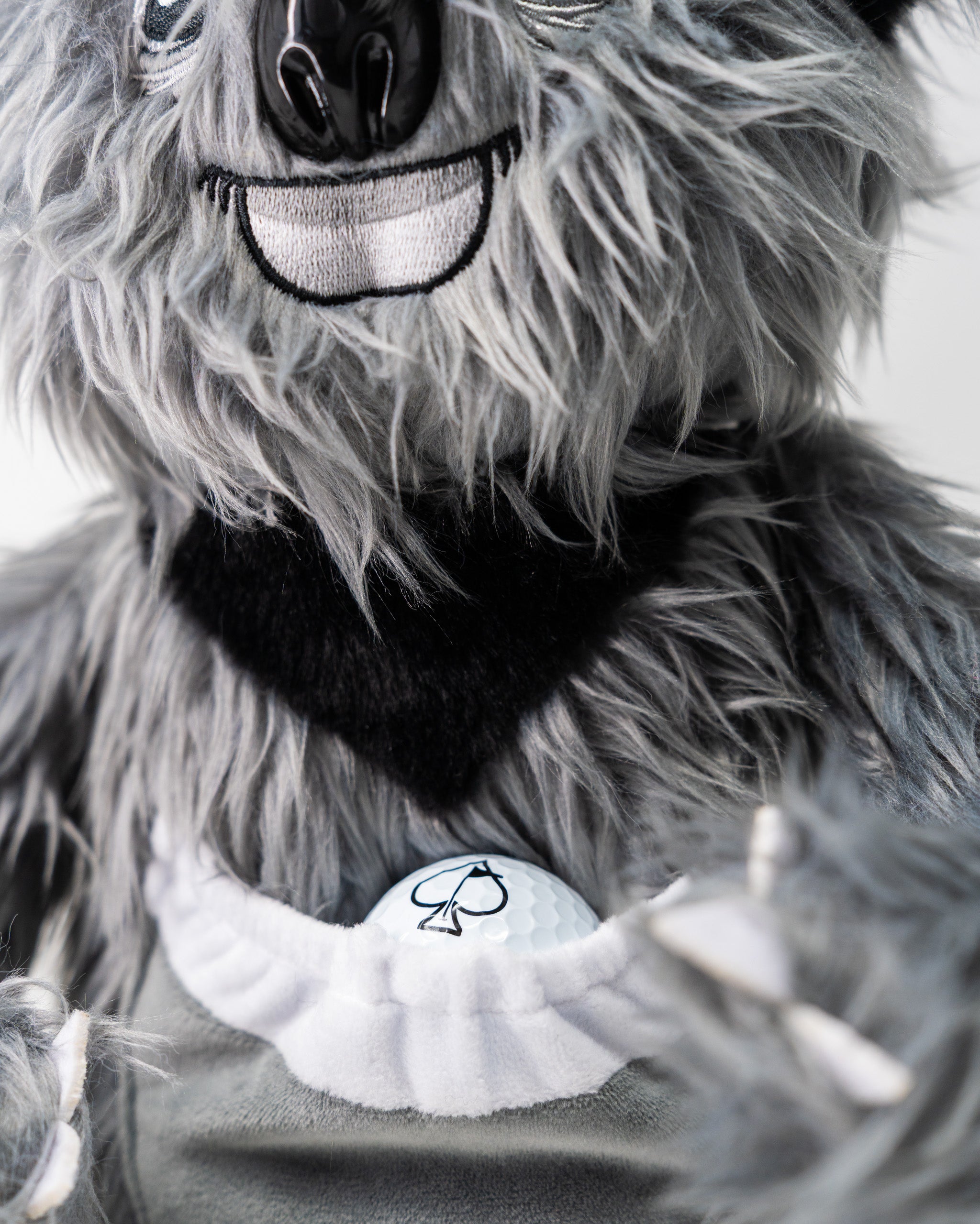 Close-up of a furry koala-themed driver cover with a smiling face, black nose, and black bandana, holding a golf ball.
