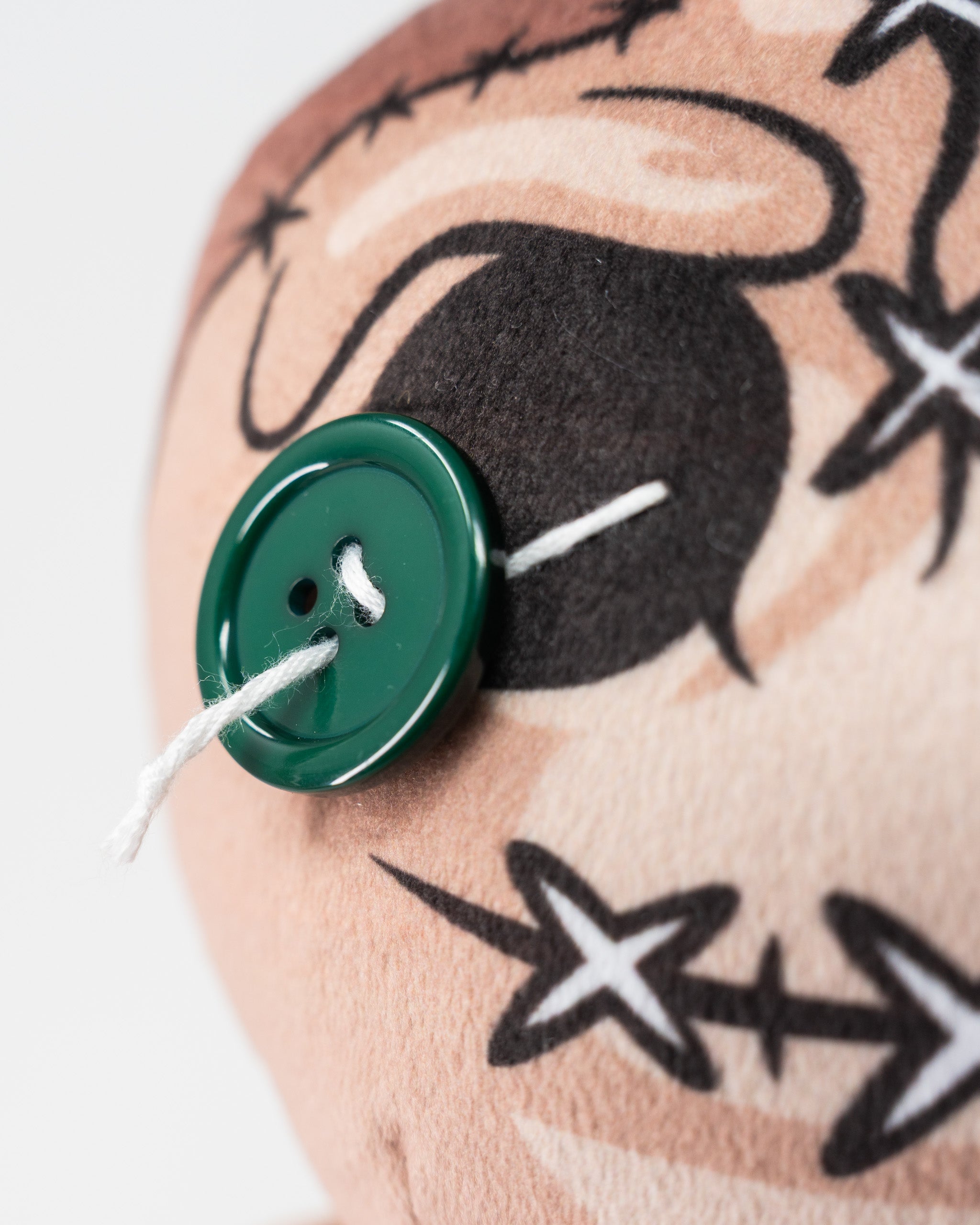 Close-up of a plush voodoo doll with a green button eye, black stitched facial features, and a white thread nose.