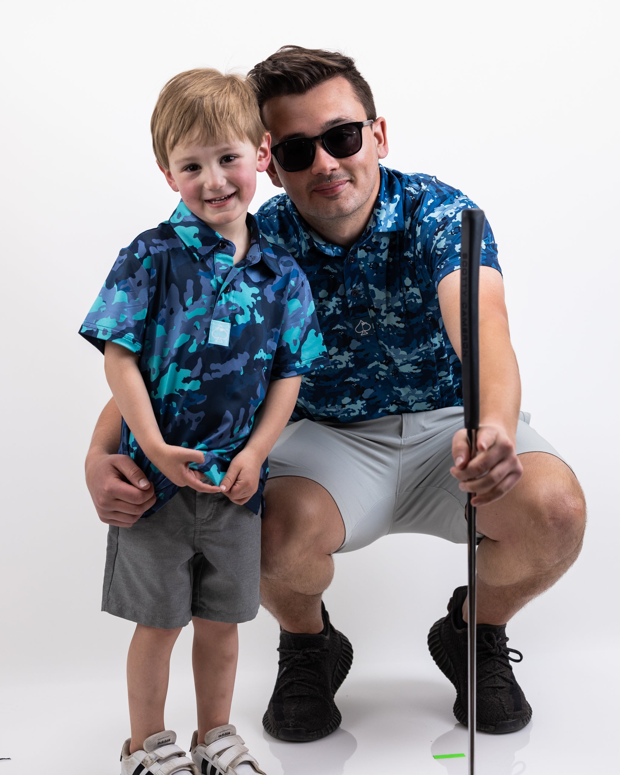 A young boy and man wearing matching blue camo golf shirts, with the man crouching and holding a golf club for a selfie.