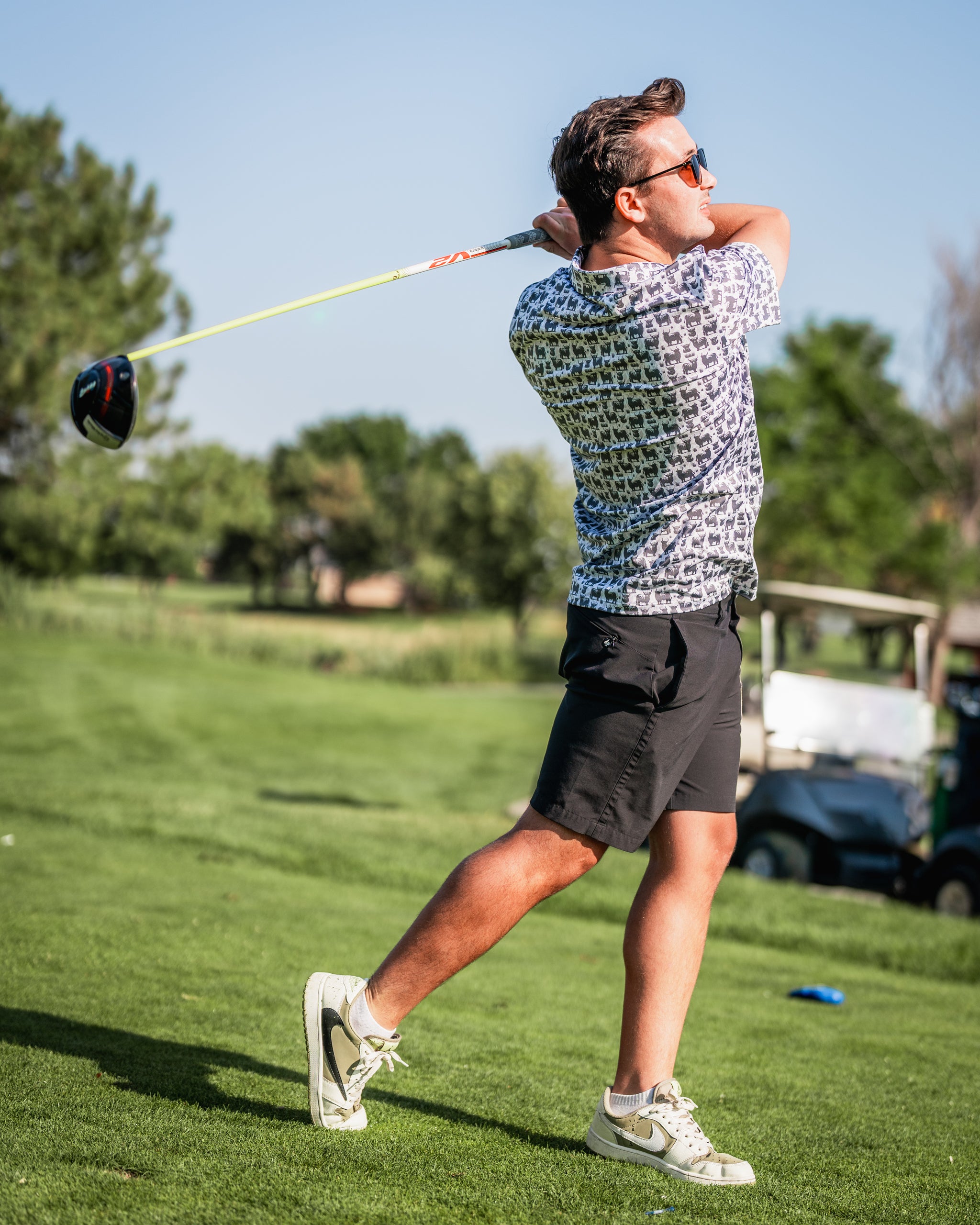 A golfer swings a driver on the course, wearing a patterned shirt and shorts, with a sunny backdrop of trees and a golf cart.