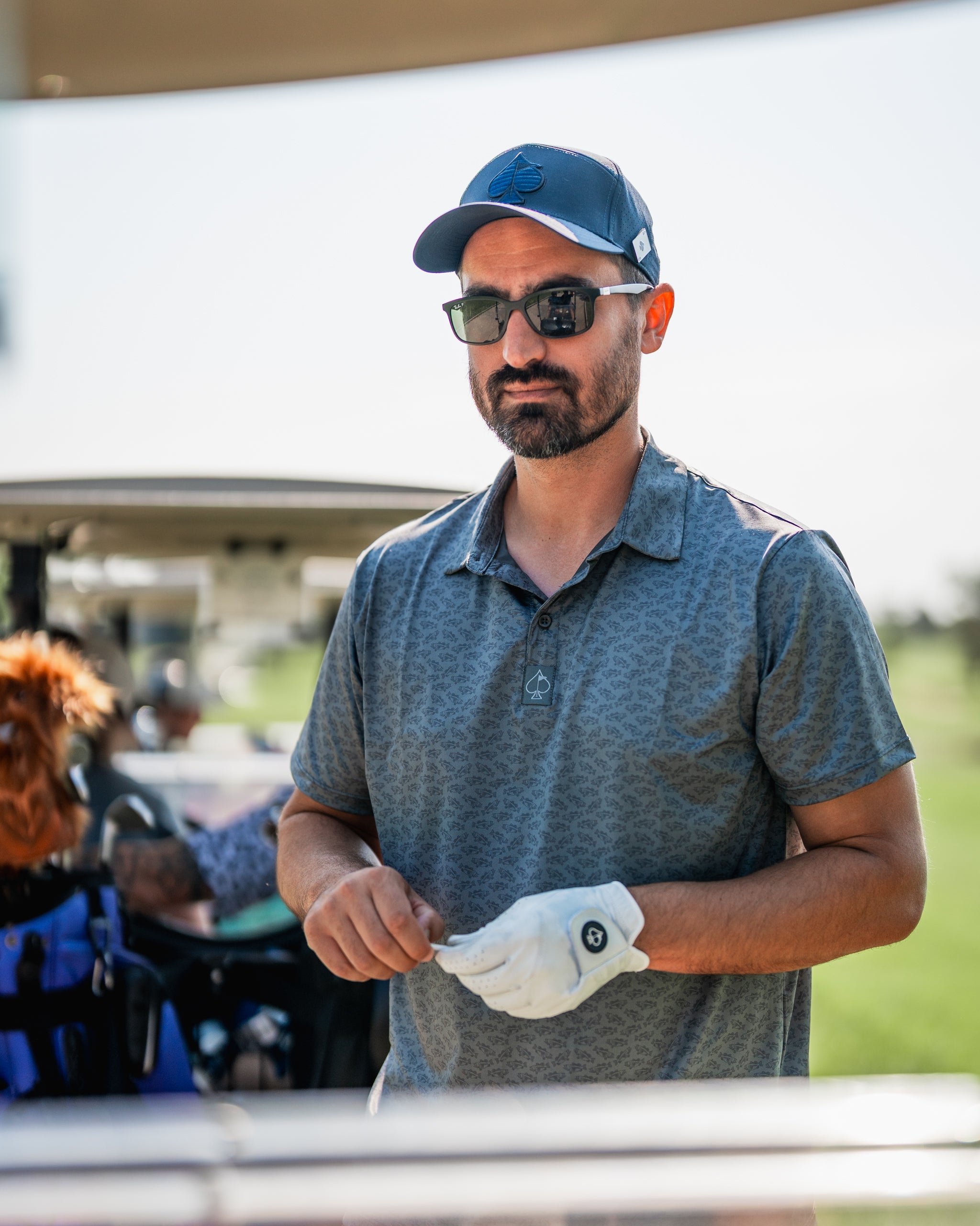 Man wearing a stylish golf shirt and cap stands by a golf cart, ready to play on a sunny day.