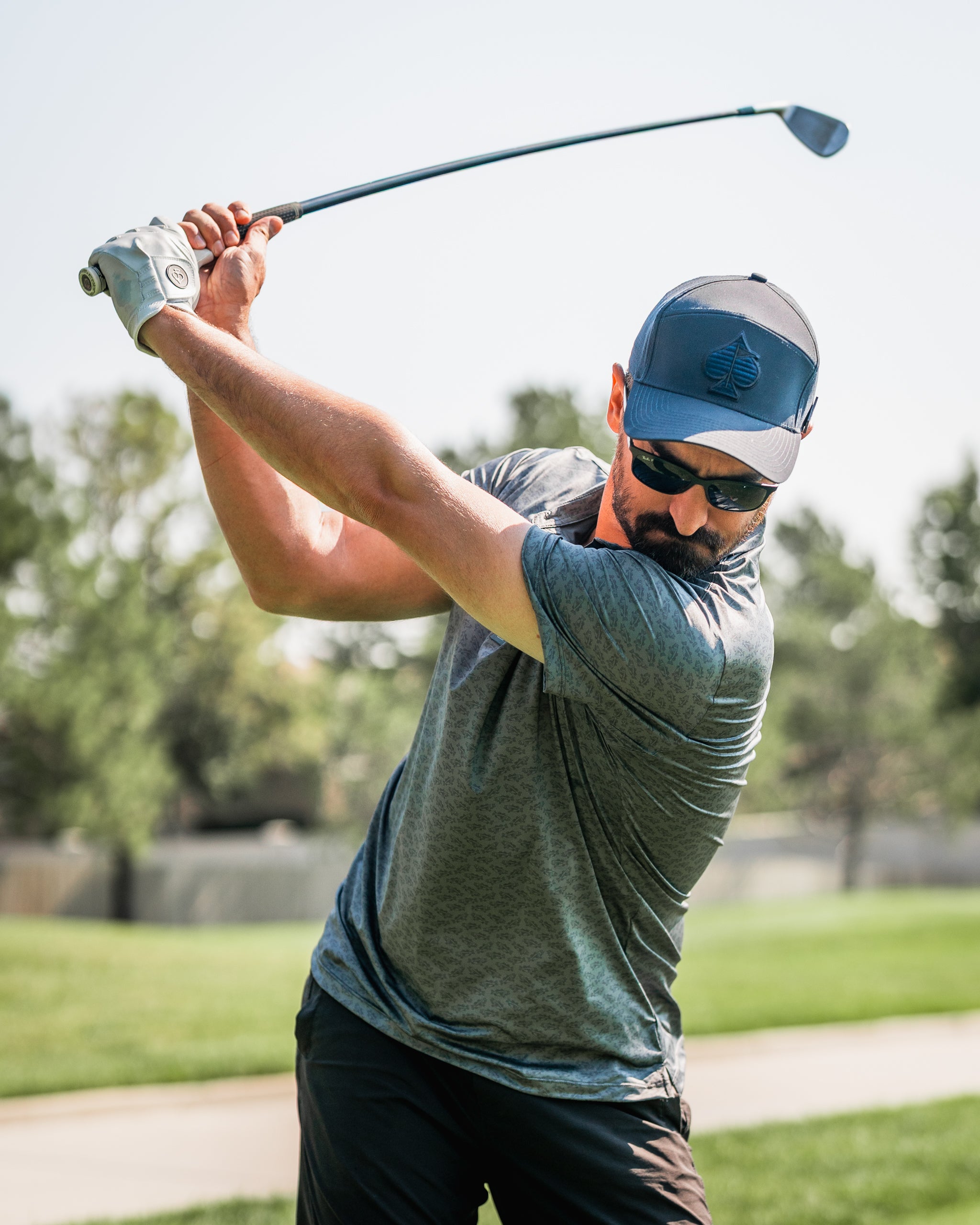Golfer in a blue cap and sunglasses swings a club on a sunny course, showcasing bold golf apparel and style.