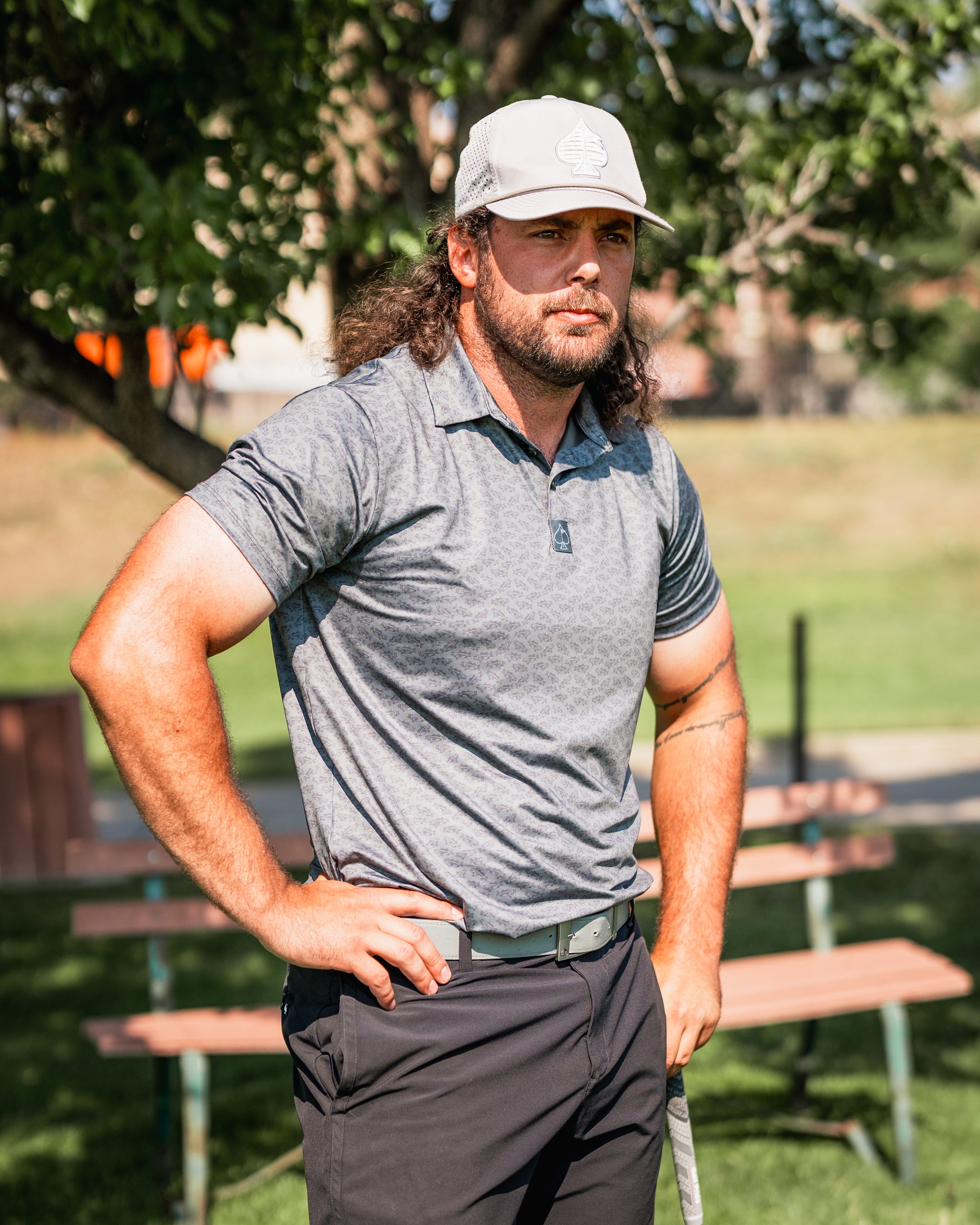 A golfer stands confidently on the course, wearing a gray polo shirt and a light cap, showcasing bold golf apparel.
