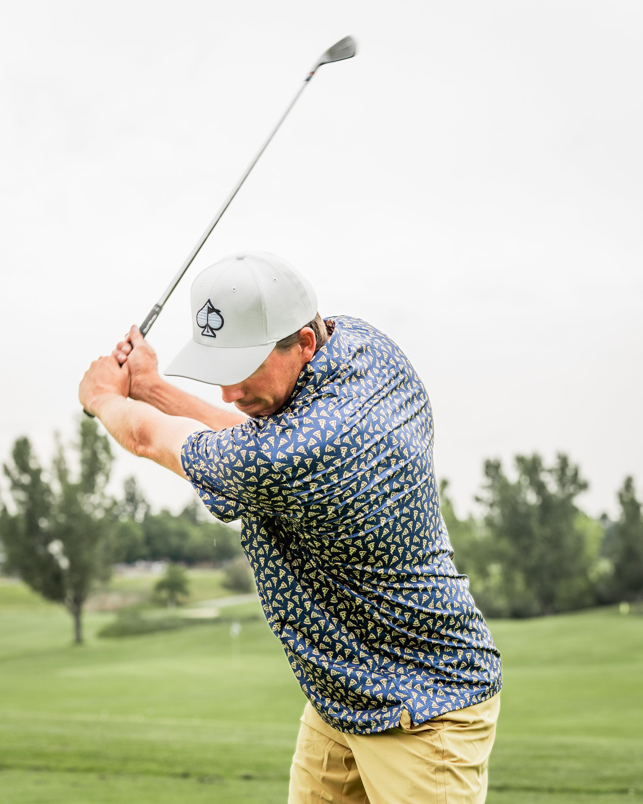 Golfer in a patterned shirt and cap swings a club on a lush green course under a cloudy sky.