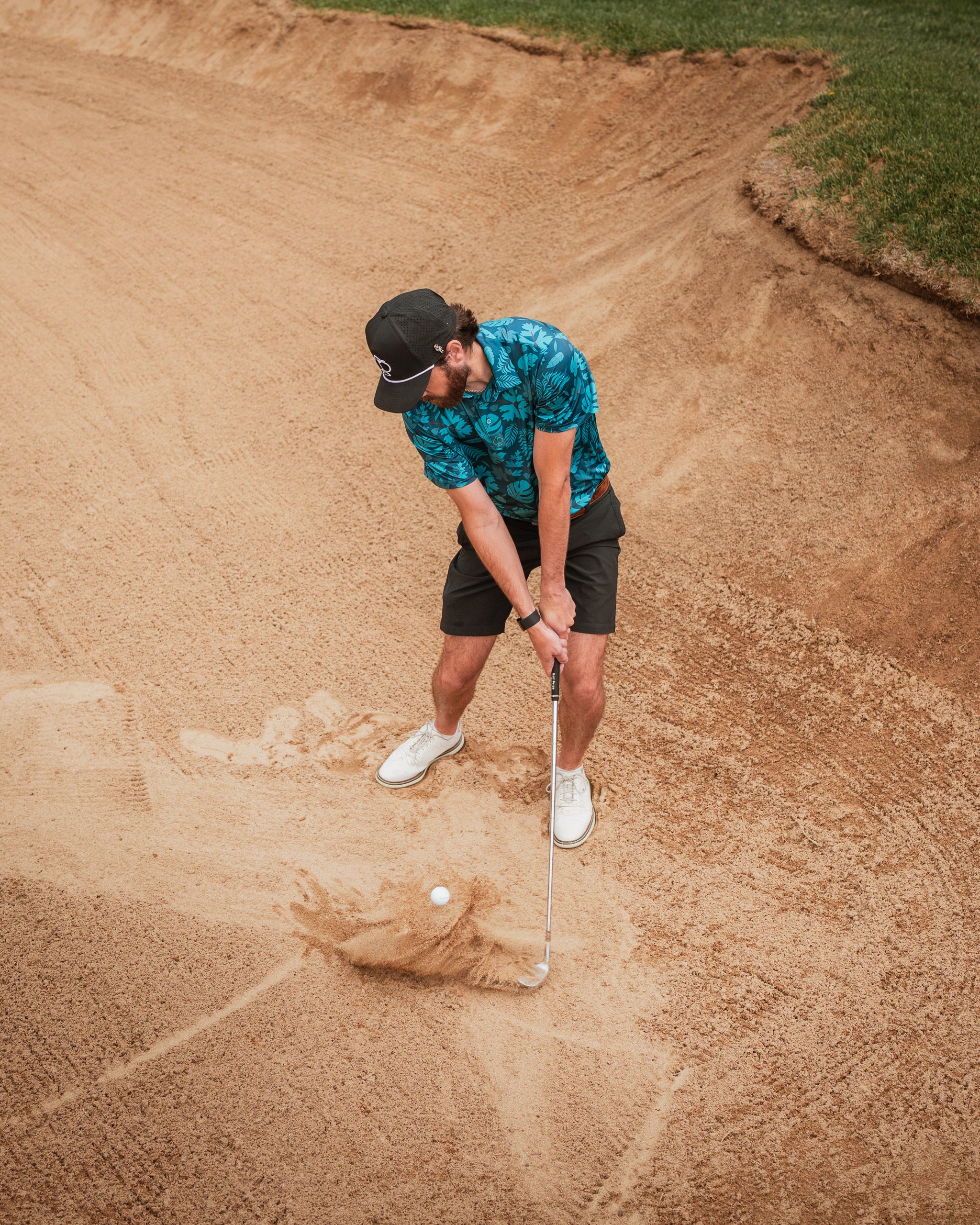 A golfer in a vibrant floral shirt and shorts prepares to hit a ball from a sand trap on the course.
